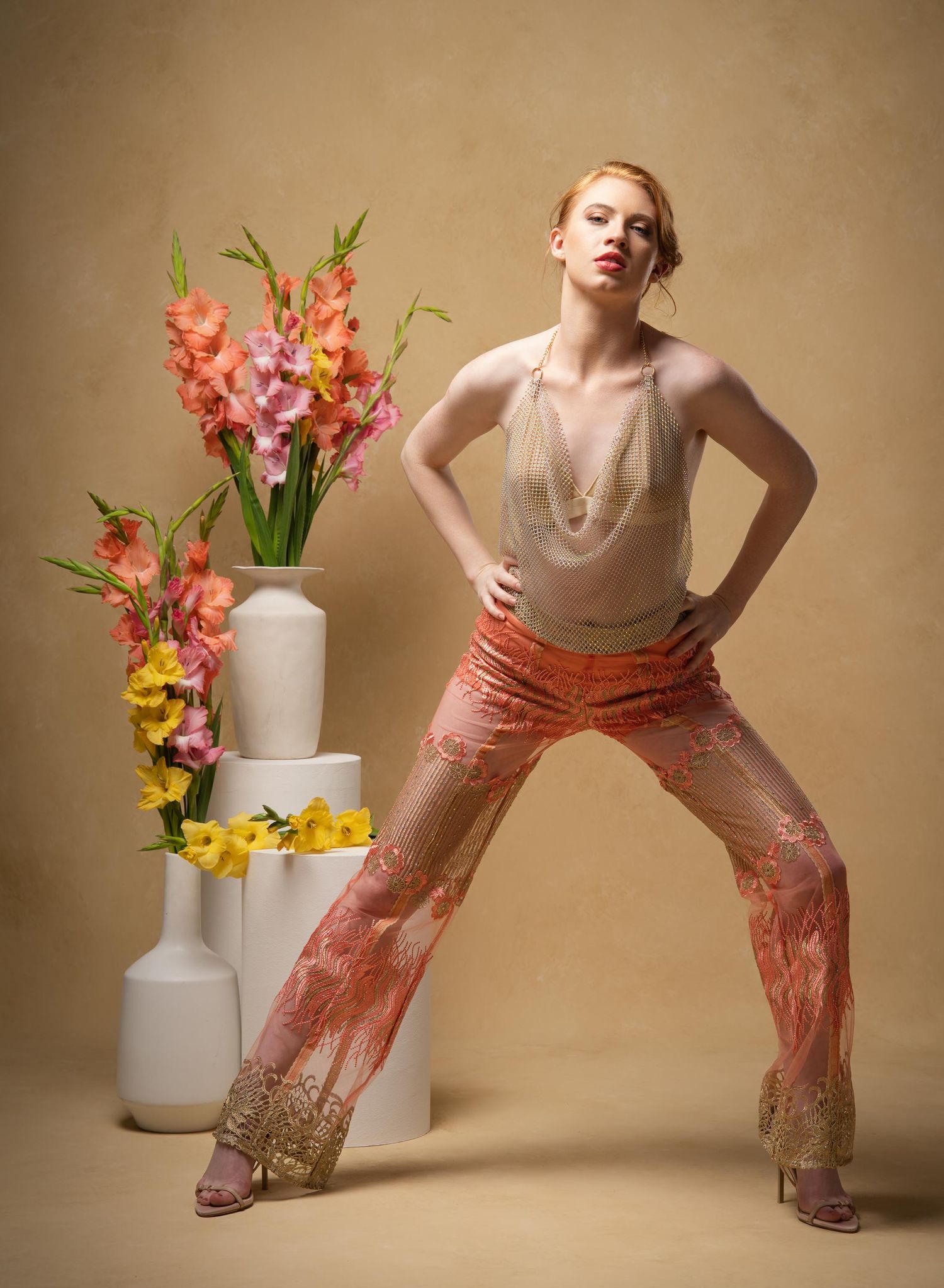Fashion photo with gladiolus flowers in white vases and model in peach floral pants against beige backdrop.