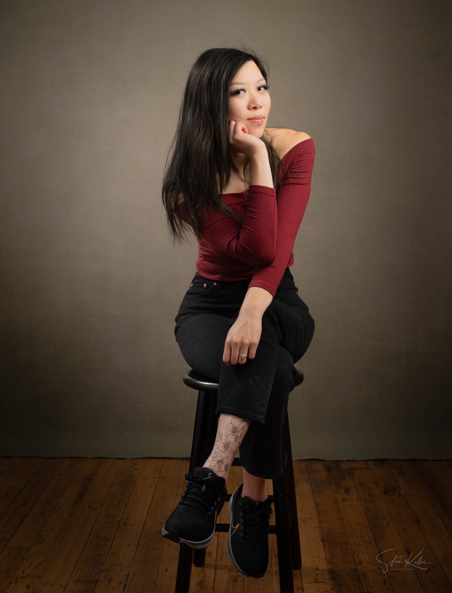 A woman in a burgundy long sleeve top and black skirt poses gracefully on a stool against a neutral backdrop.