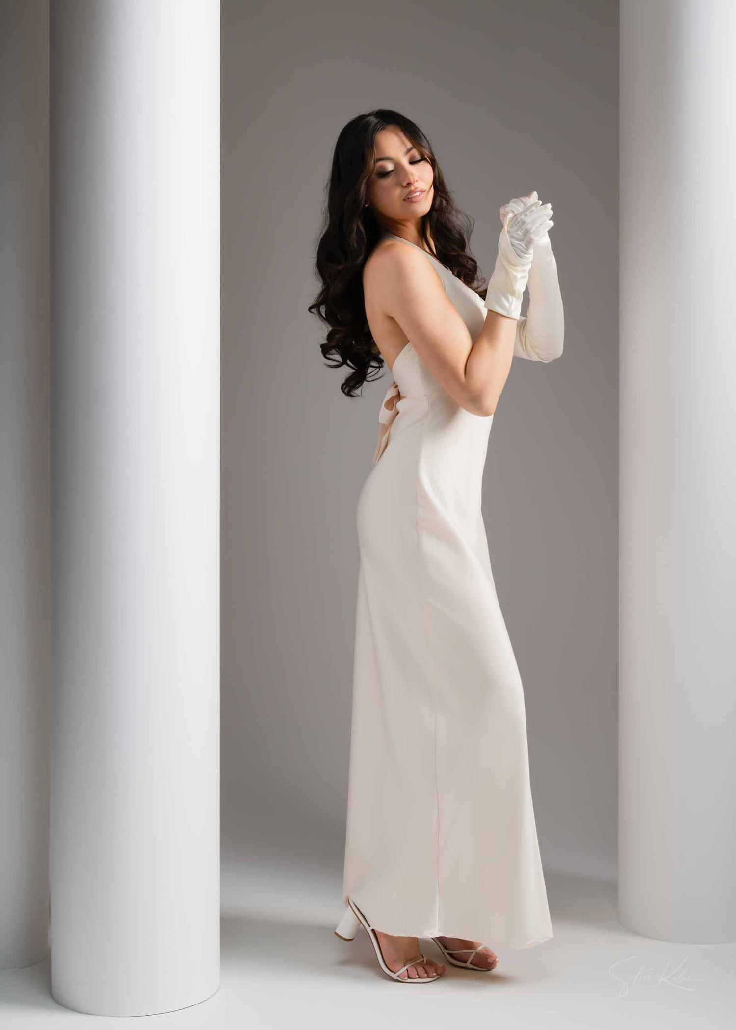 A person in a white slip dress poses elegantly against white columns in a minimalist studio setting.