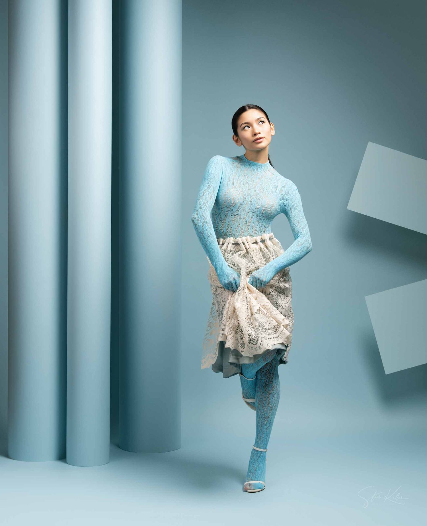 A figure in a light blue sweater and floral skirt poses gracefully against a minimalist blue-toned studio backdrop.