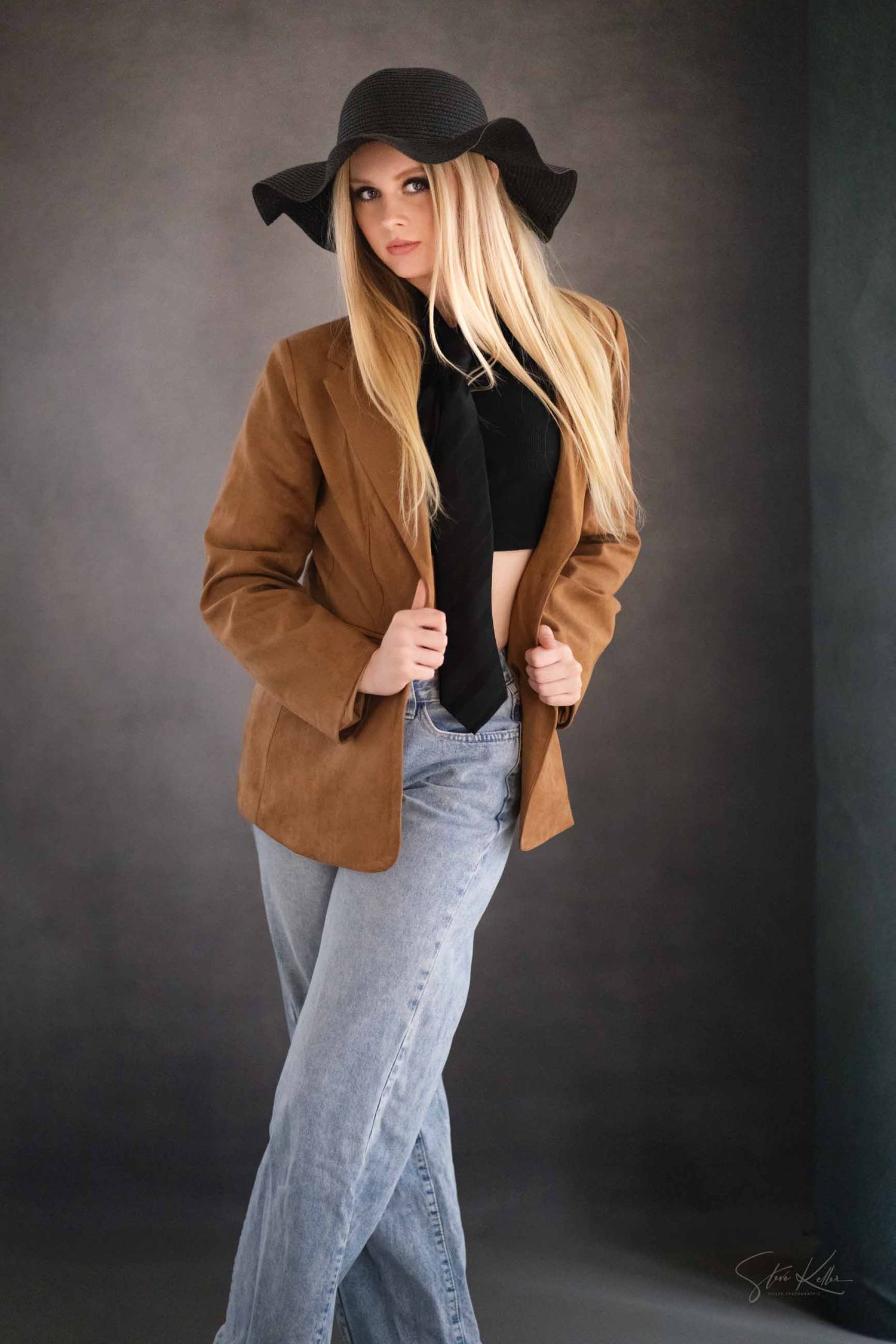 Fashion portrait featuring a brown jacket, black hat, and acid wash jeans against a dark studio background.