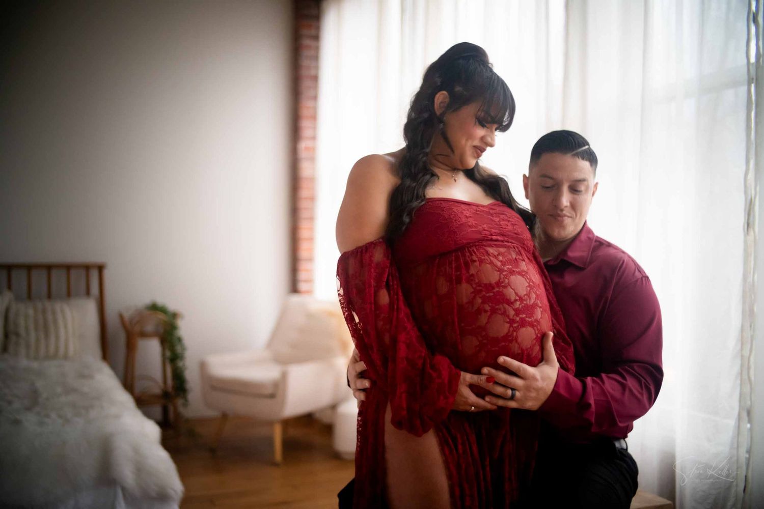 A couple in burgundy and maroon outfits embrace in front of bright windows in a minimalist room setting.