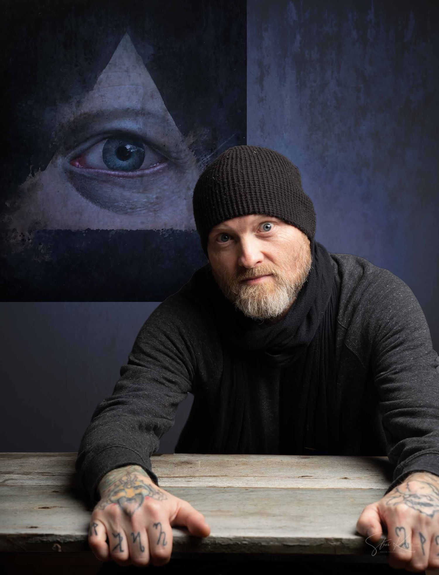 A person wearing a black beanie and coat leans on a weathered wooden surface in moody blue lighting.