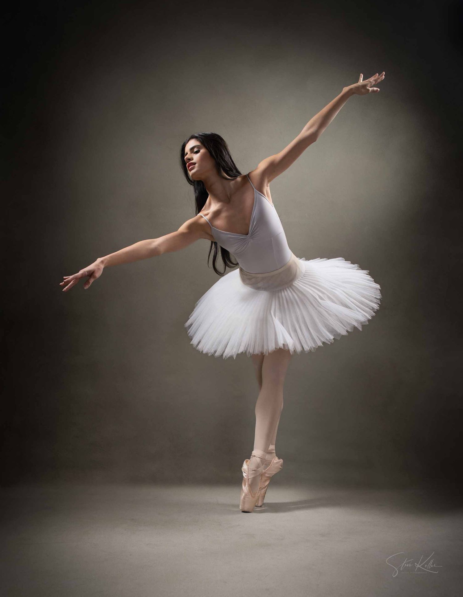 A ballet dancer in a white tutu performs an elegant arabesque pose against a dark studio backdrop.