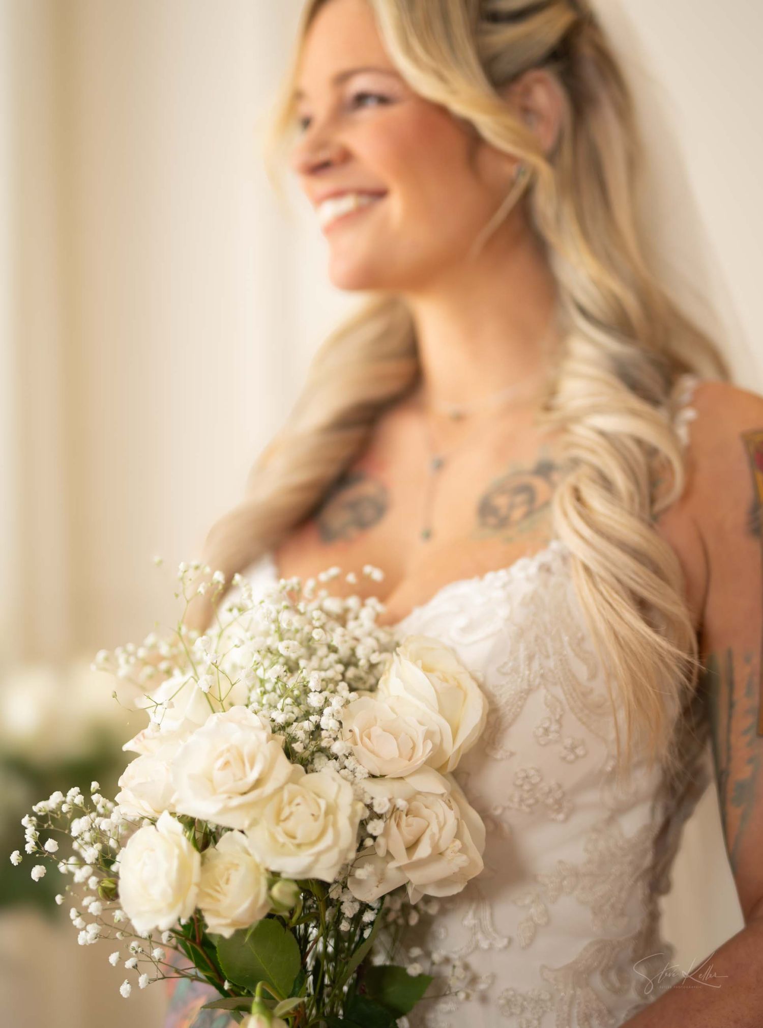 A bride in a white lace dress holds a bouquet of white roses and baby's breath in a soft natural light setting.