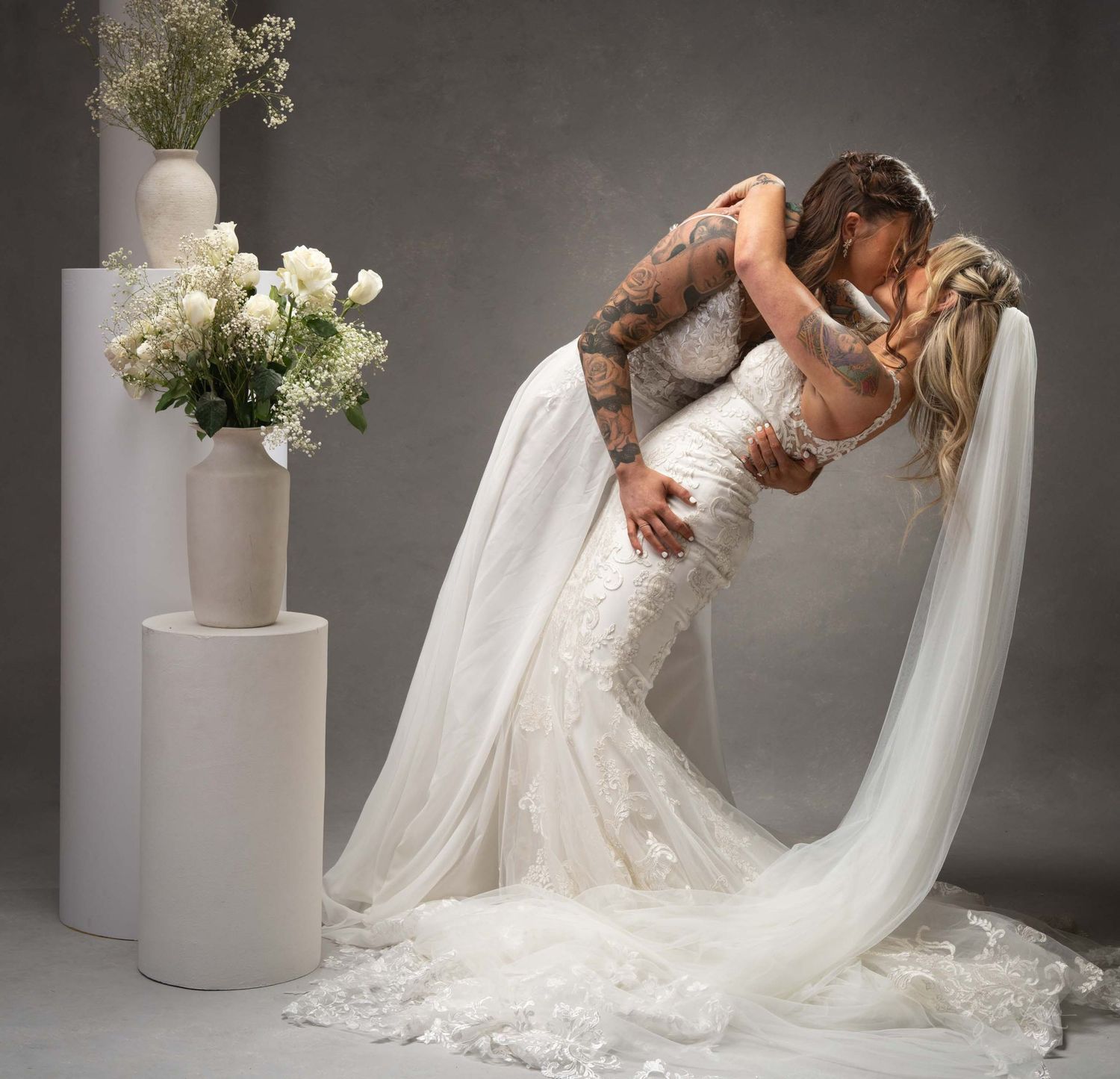 A romantic wedding photo of a couple in white embracing in front of floral arrangements on white pedestals.