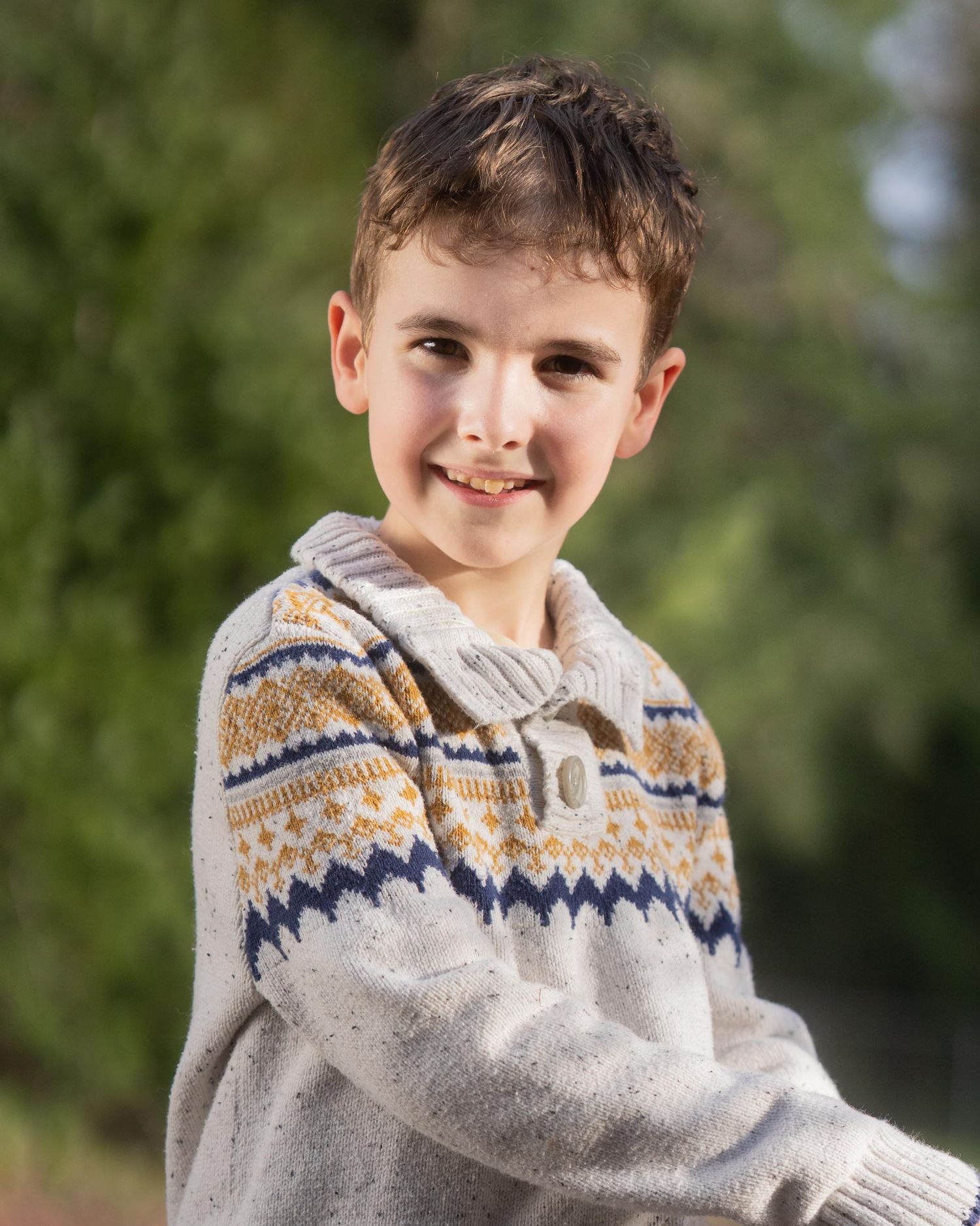 A young person in a cozy patterned sweater smiles warmly against a soft green outdoor background.