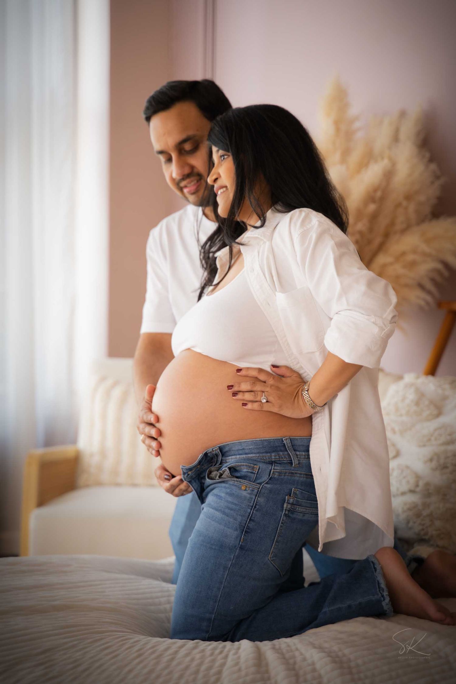 Couple in white shirts kneeling on bed, man embracing pregnant woman from behind, both smiling at her bare baby bump.