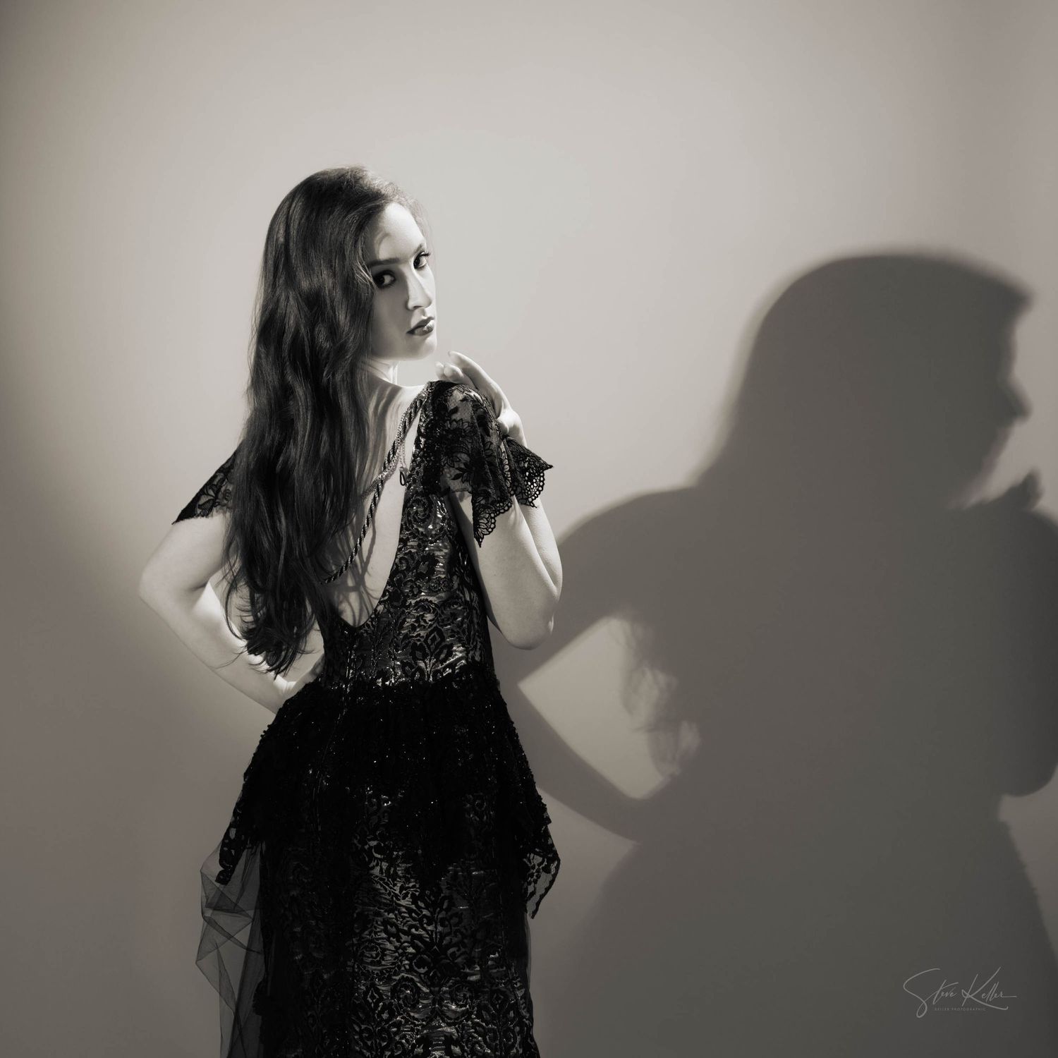 A moody black and white portrait of a model in a lacy black dress casting dramatic shadows on a wall.