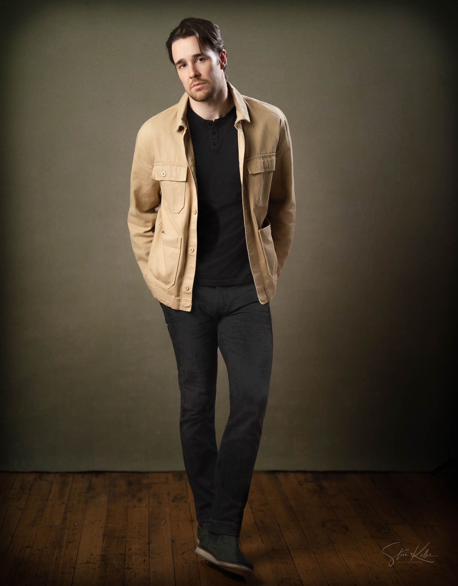 A person in a tan jacket and black pants poses in a studio setting against a dark gradient background.