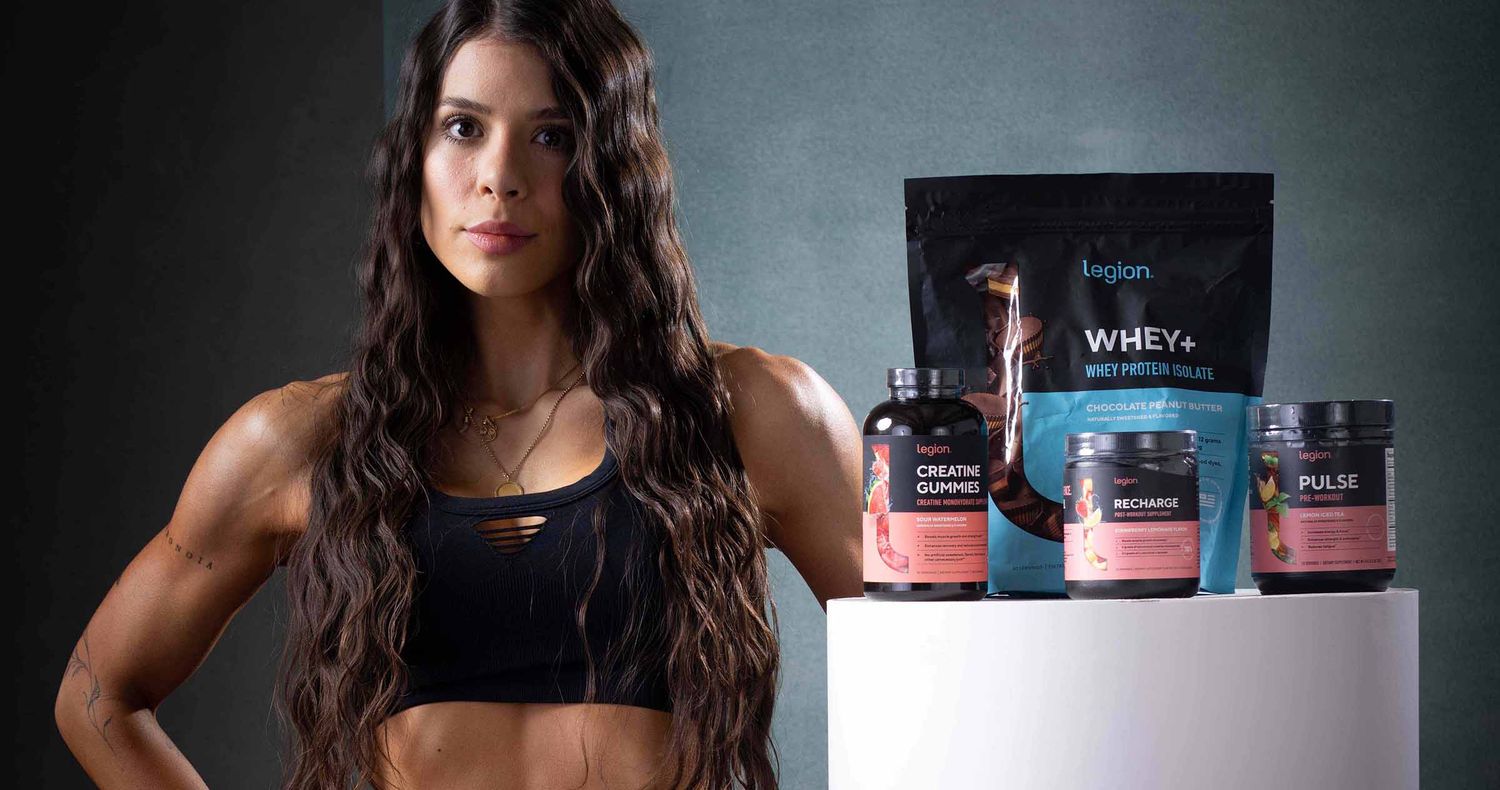 Fit woman with long wavy dark hair in a black sports top stands beside a display of whey protein, creatine, and pre-workout supplements.