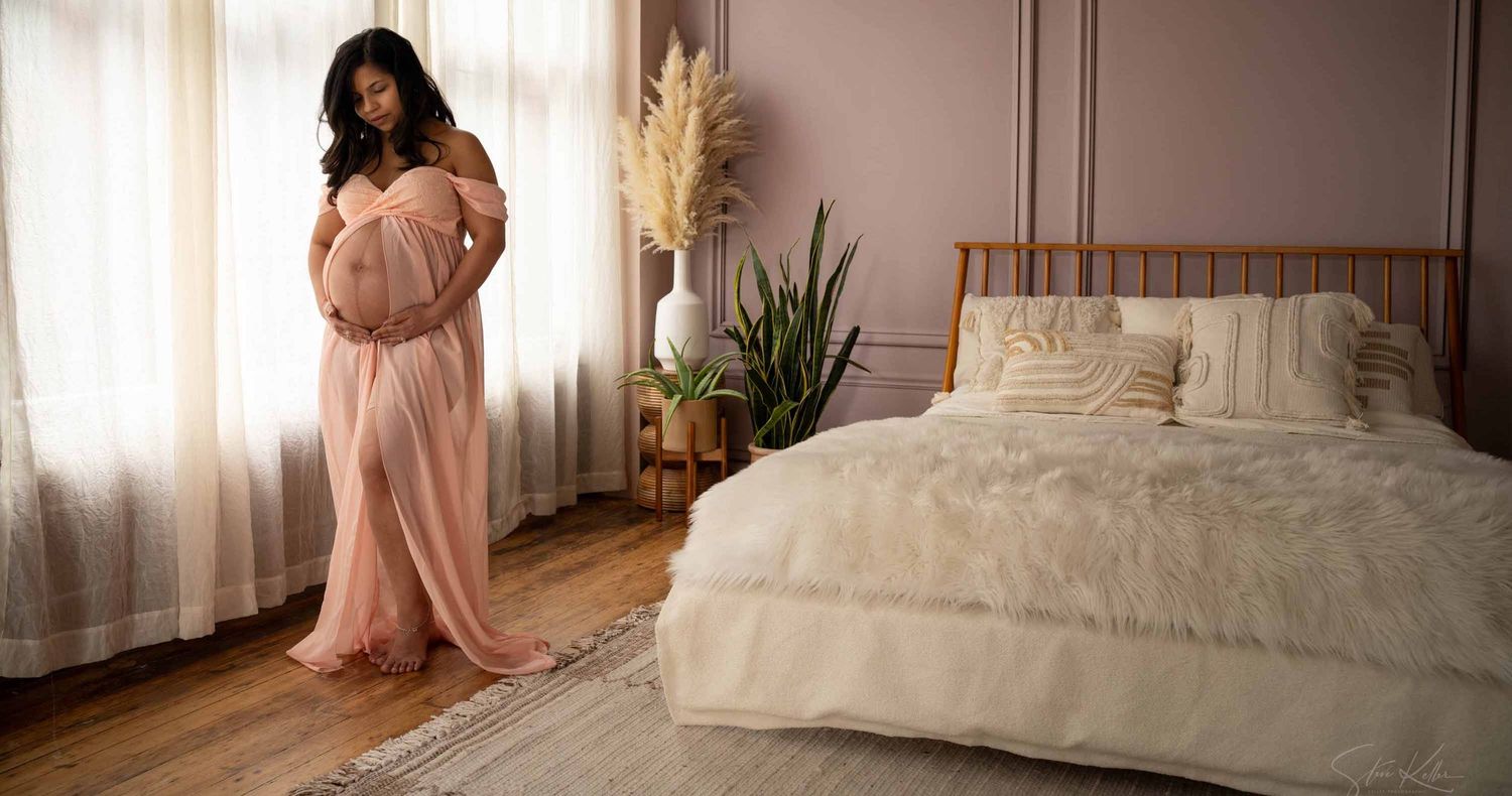 Pregnant woman in a flowing blush pink maternity gown cradles her belly beside a cozy bed in a softly lit mauve bedroom.