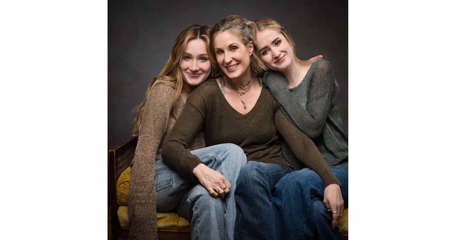 Three smiling women in casual sweaters and jeans pose close together in a warm studio portrait against a dark backdrop.