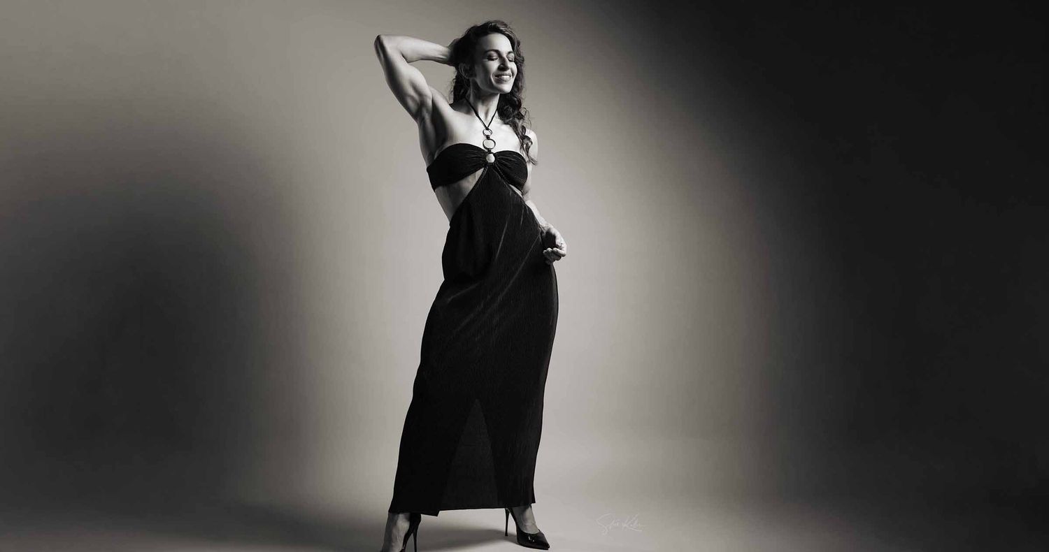 Black and white studio portrait of a woman posing in a long black cutout dress and heels, one arm raised above her head.