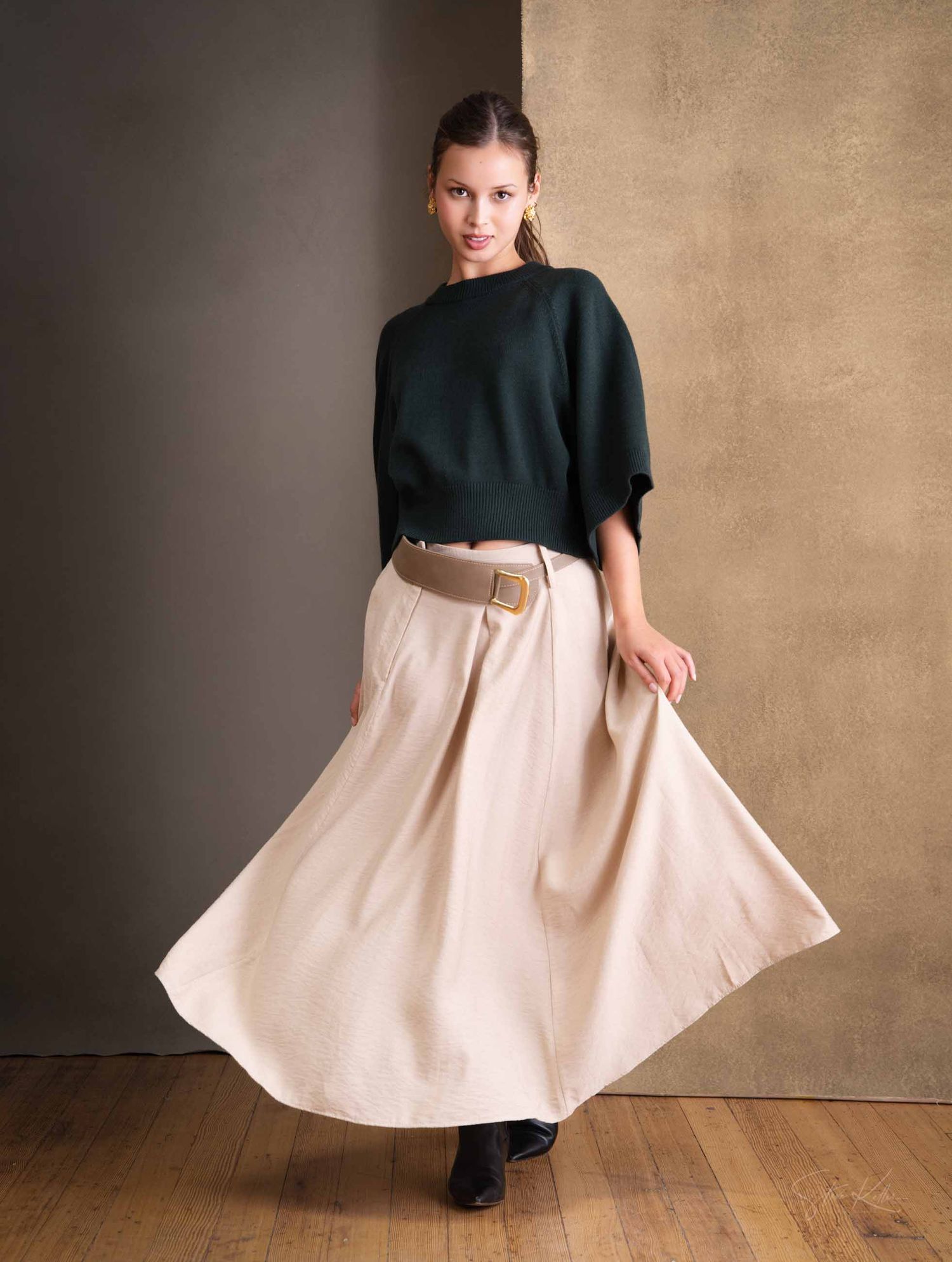 Fashion model wearing a dark oversized sweater with cream maxi skirt in studio setting with beige backdrop and wooden floor.