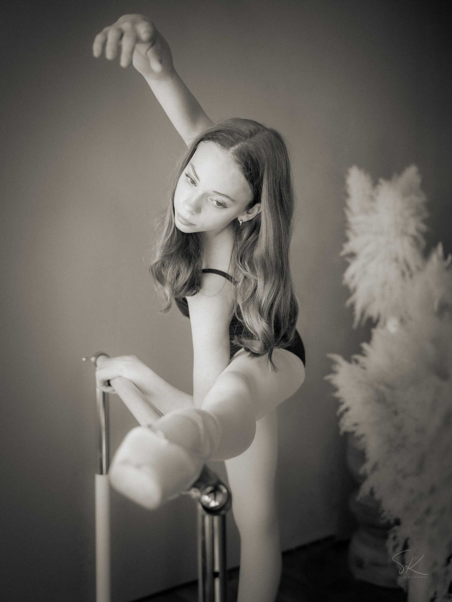 Black and white photo of a young dancer gracefully posing at a ballet barre with one arm raised.