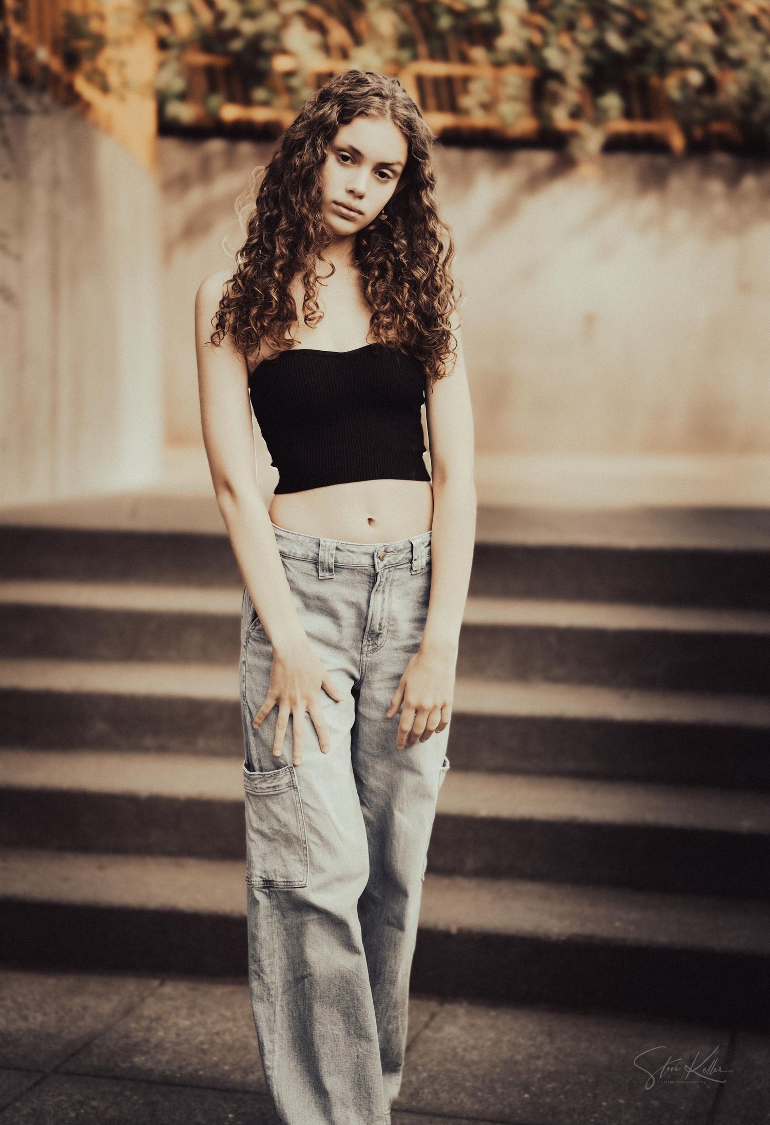 A person in a black crop top and baggy cargo pants poses on concrete steps in a warm-toned portrait.