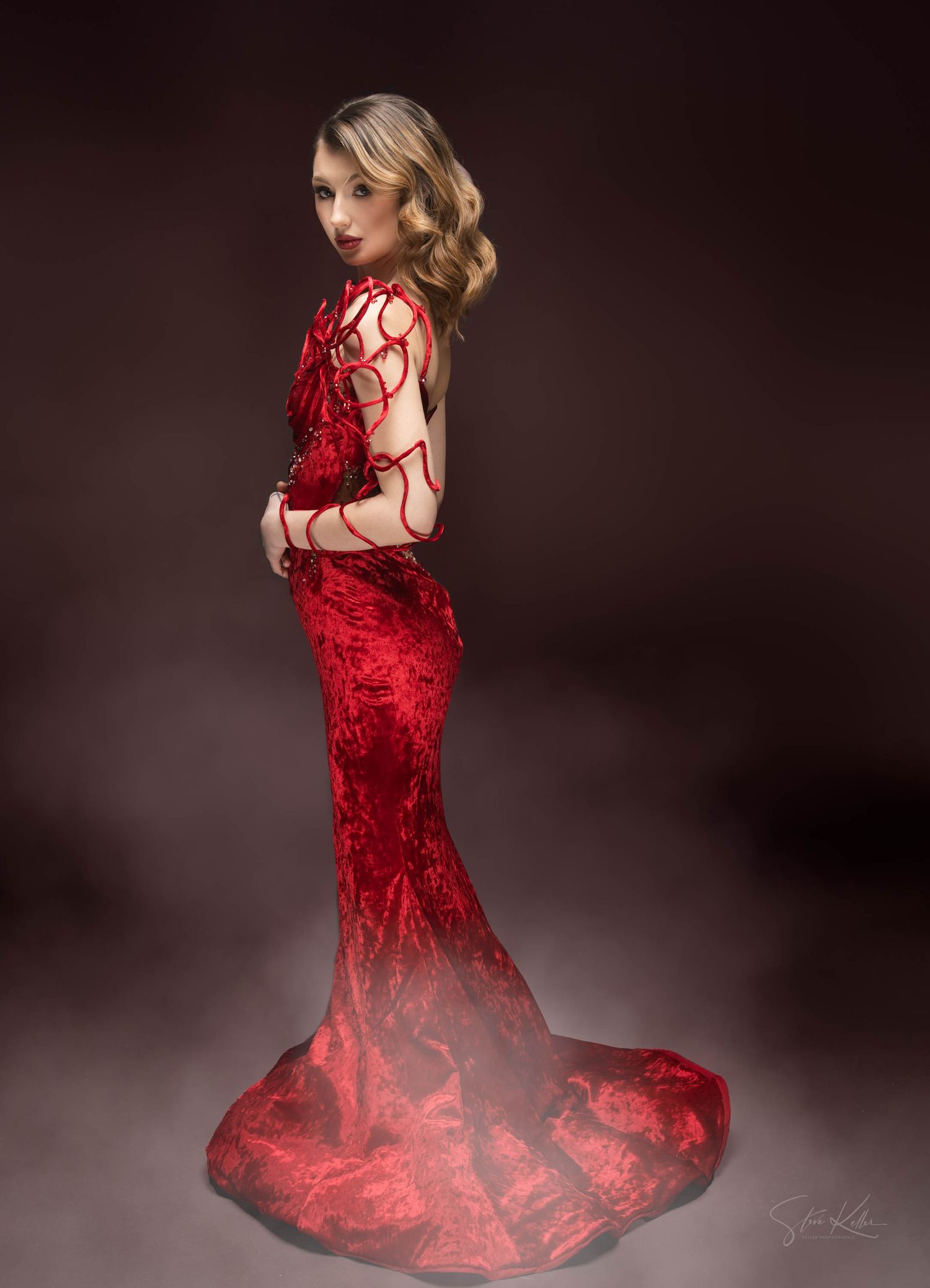 Model showcases a glamorous red lace gown with mermaid silhouette against a dark studio backdrop.