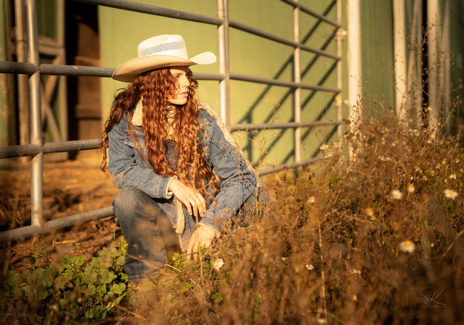A woman with long curly red hair wearing a cowboy hat and denim jacket poses near a rustic fence in a dry field.