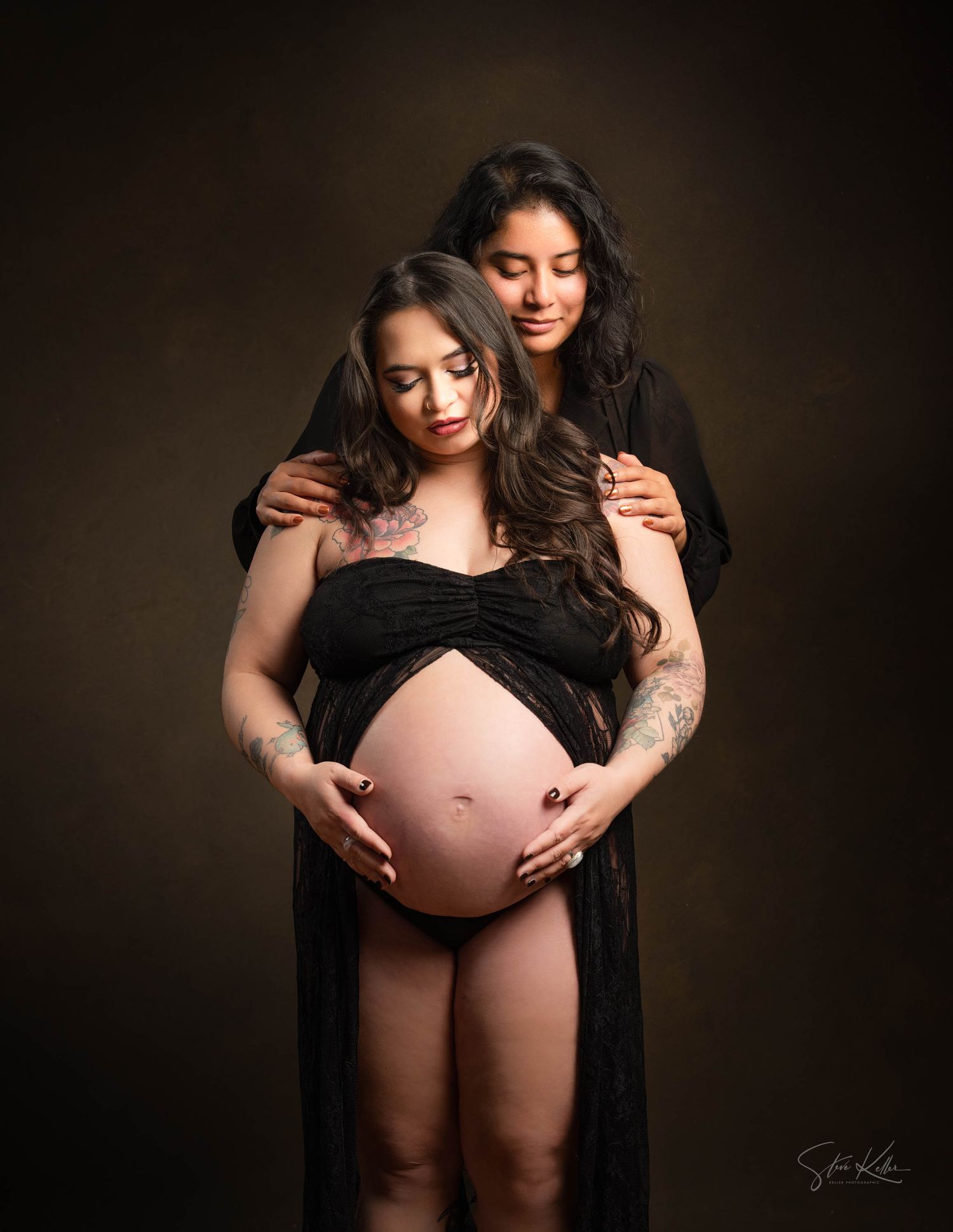 Intimate maternity portrait with two figures in black dresses against dark background.