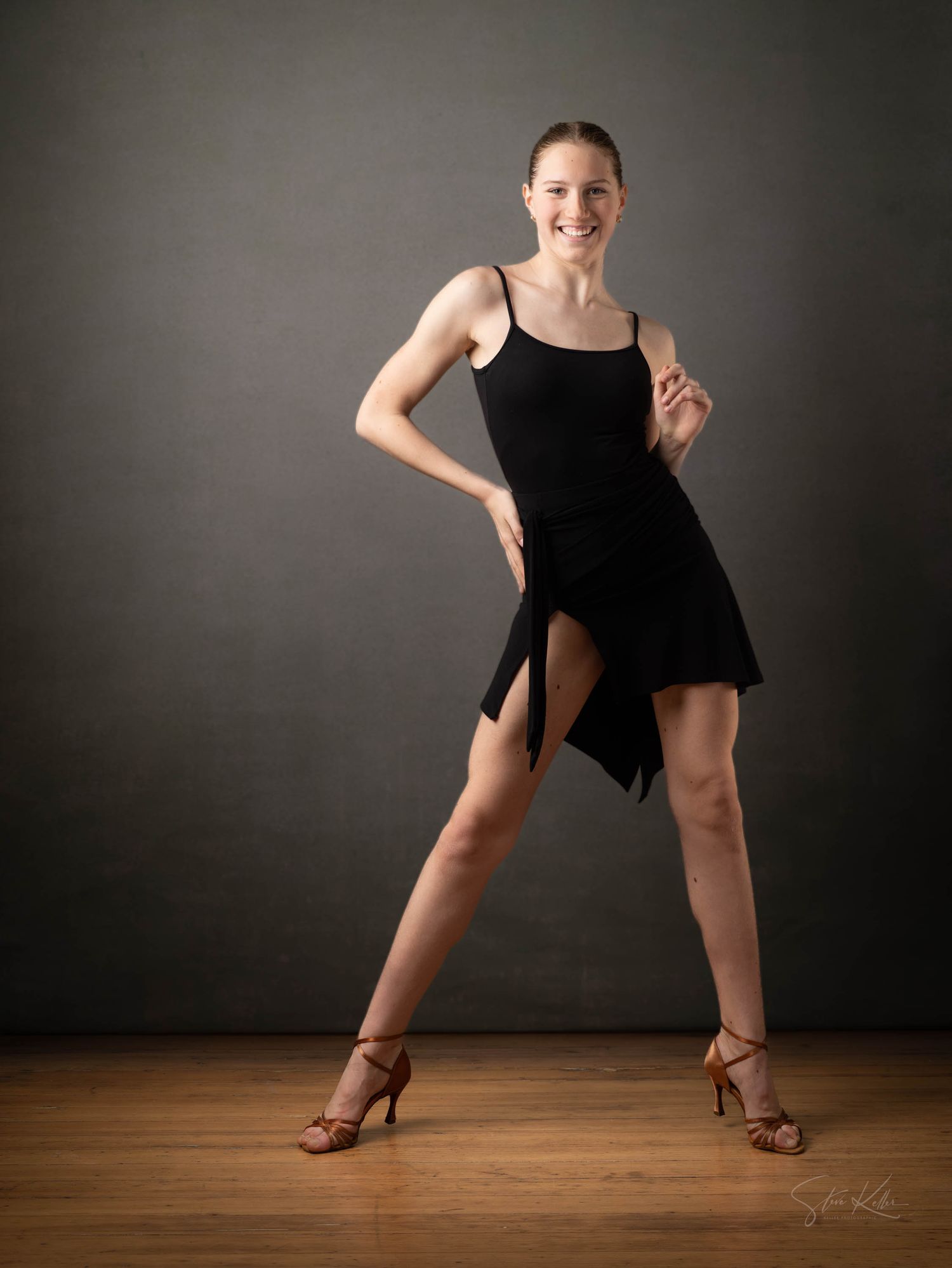 A dancer in a black dress strikes an energetic pose with one leg bent, performing on a wooden floor against a dark background.