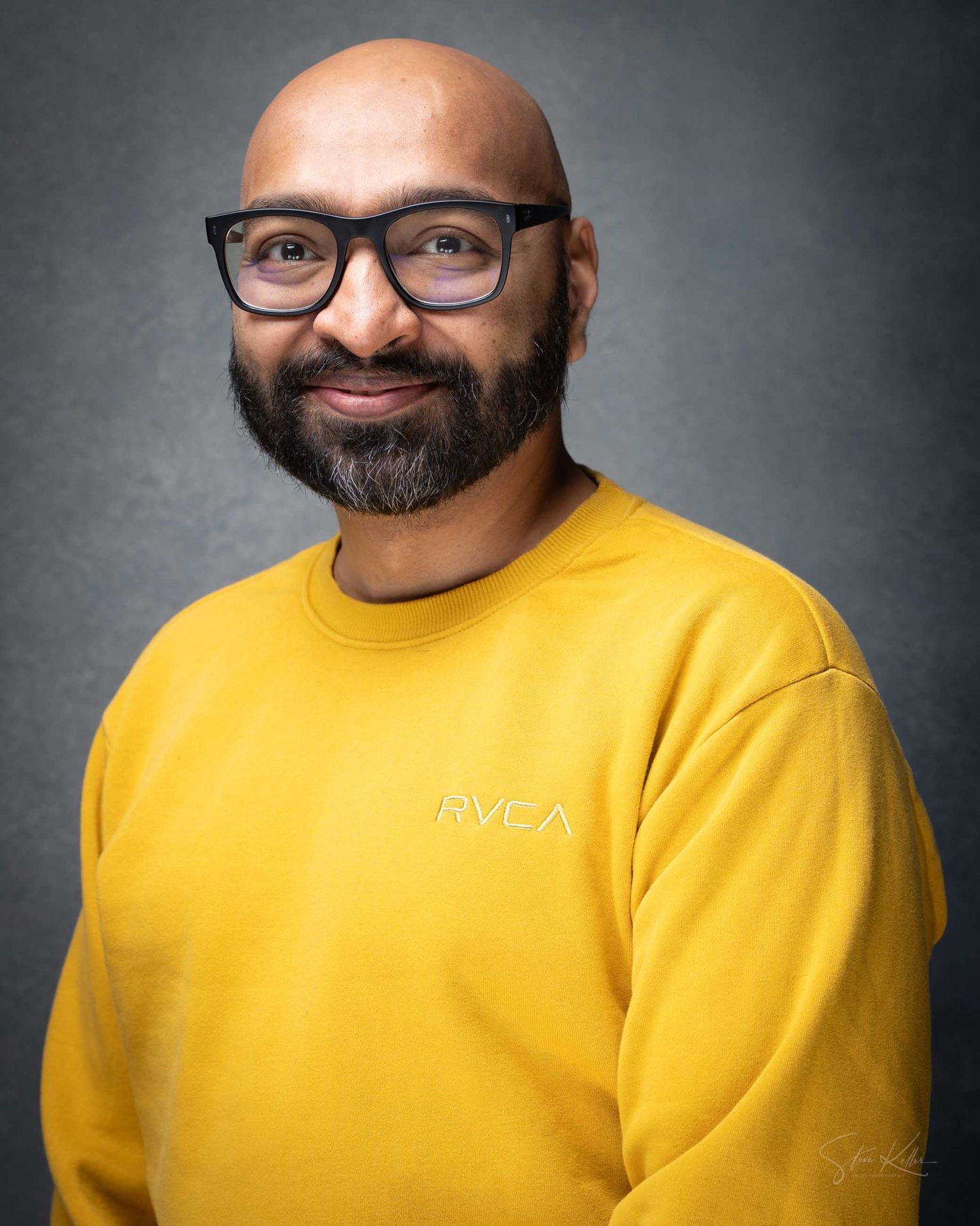 Bald man with beard and glasses wearing a yellow RVCA sweatshirt, smiling for a professional headshot.