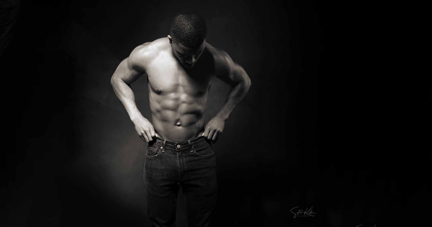 Shirtless muscular man with defined abs looking down while gripping the waistband of his jeans in a dramatic low-key dark studio portrait.