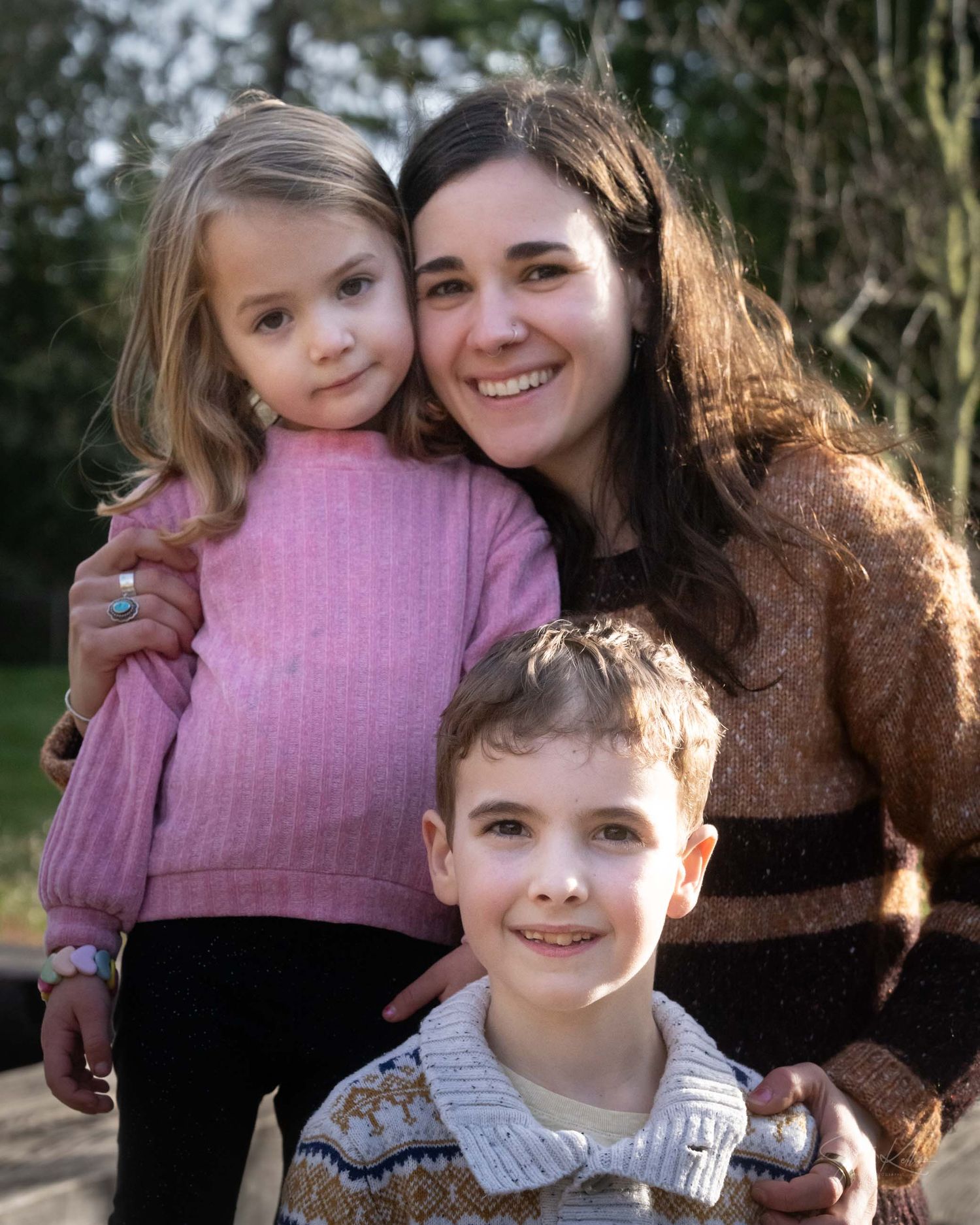 A smiling woman poses outdoors with two young children, a girl in pink and a boy in a striped sweater.