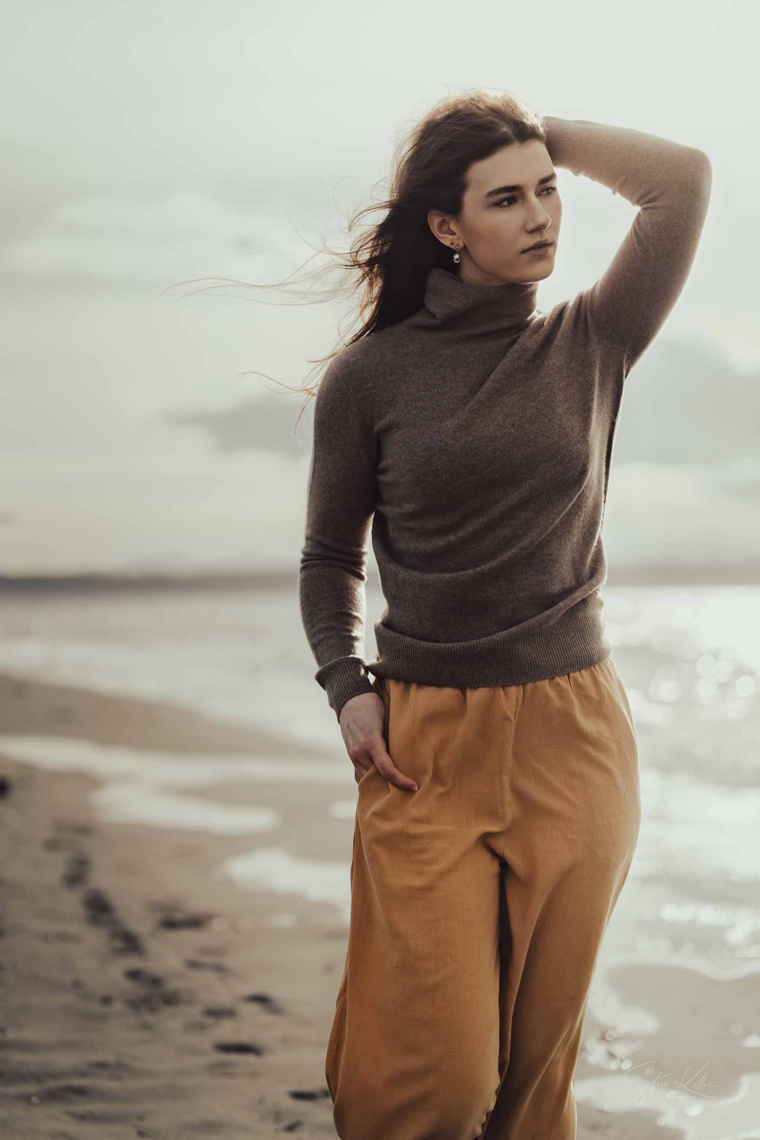 A person in a brown sweater and mustard pants stands thoughtfully on a misty beach shoreline.