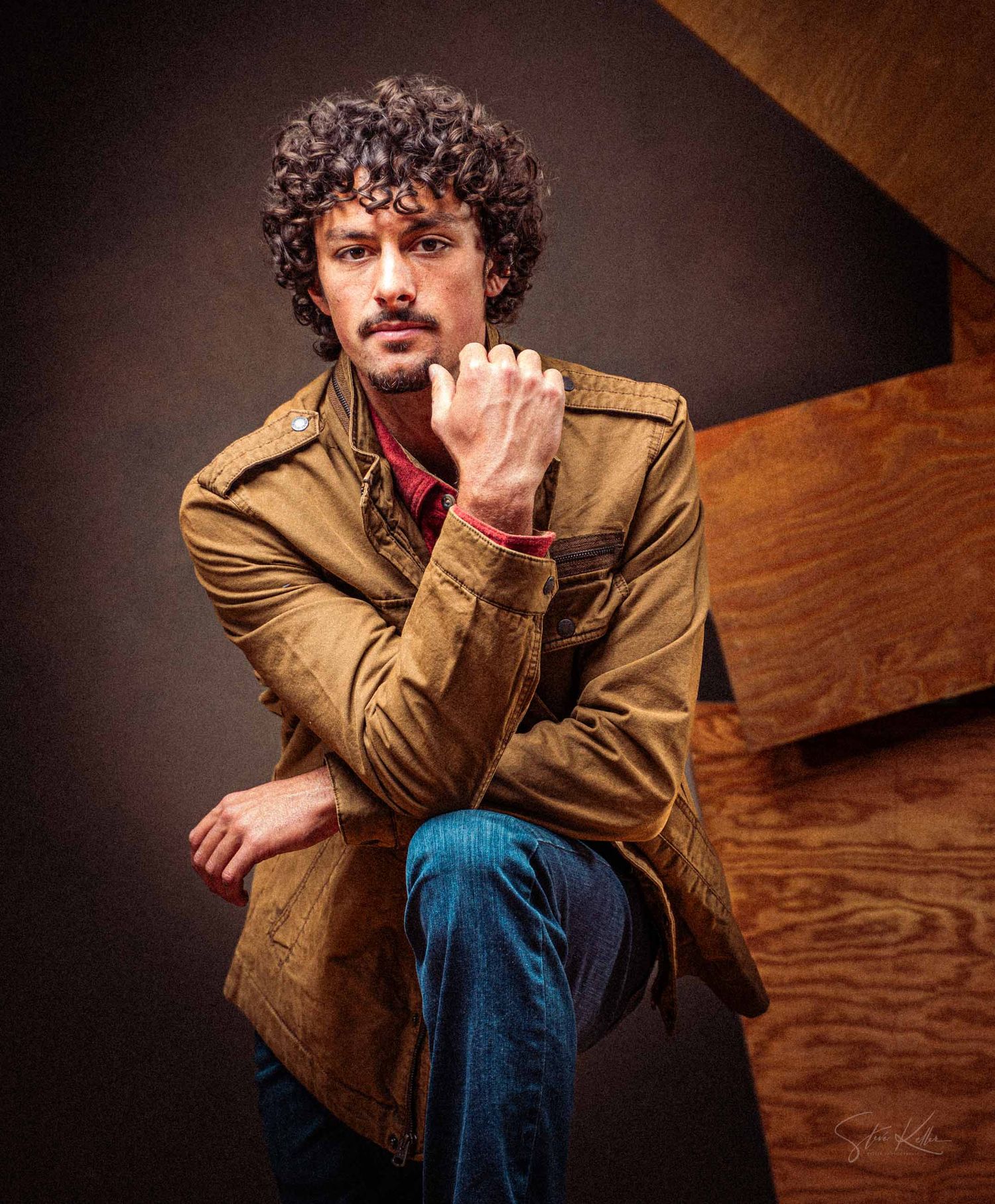 A person in a brown jacket and jeans sits pensively on wooden stairs in a dark studio setting.