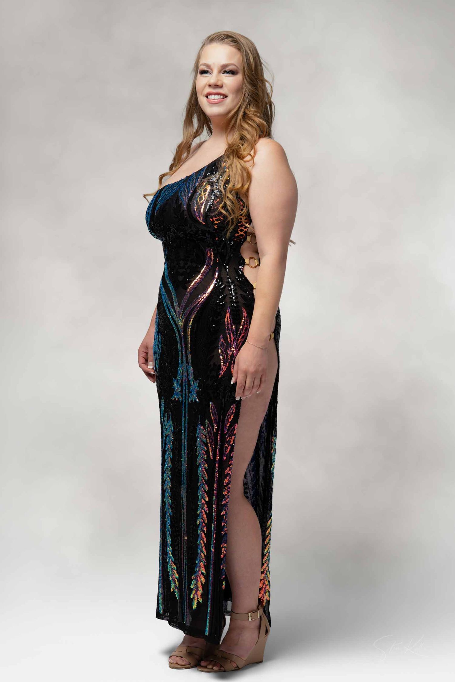 A woman in a long black velvet dress with colorful accents poses against a light gray studio backdrop.