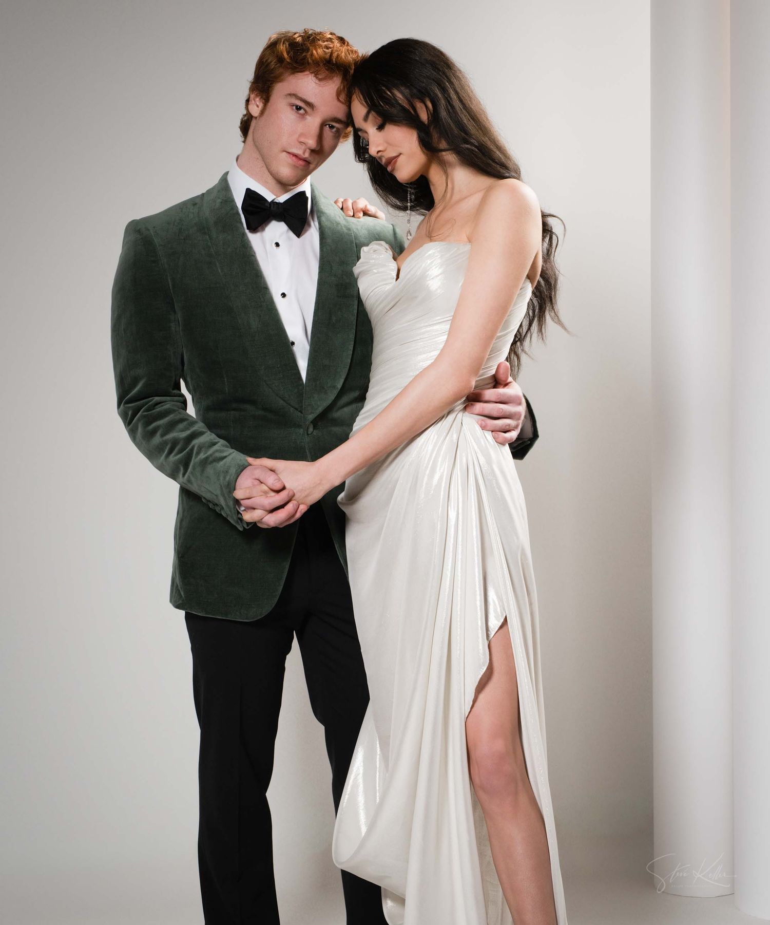 Romantic couple in formal attire pose together against white backdrop, with bride in flowing white gown and groom in tuxedo.