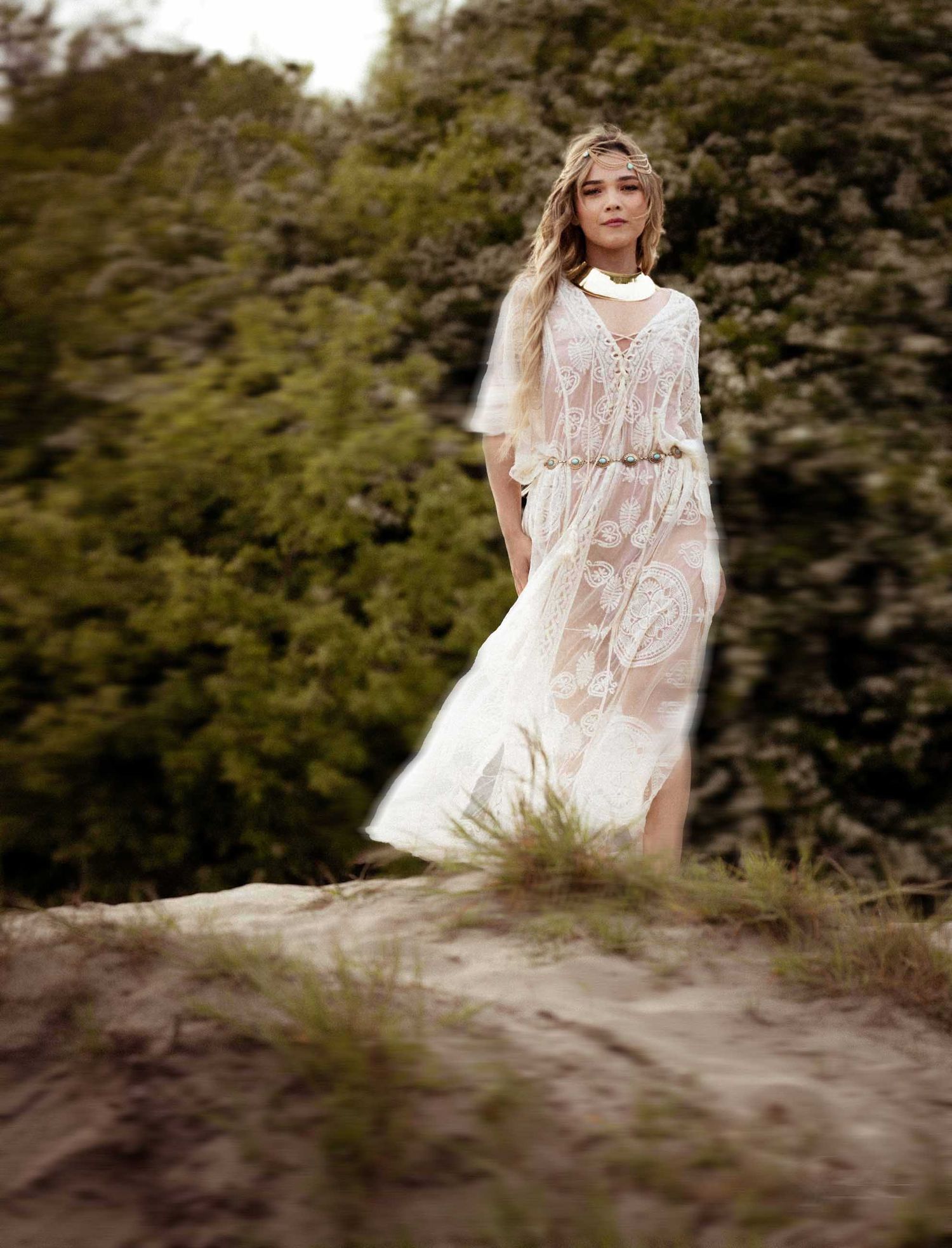 A person in a flowing white lace dress poses gracefully in a natural outdoor setting with trees in the background.