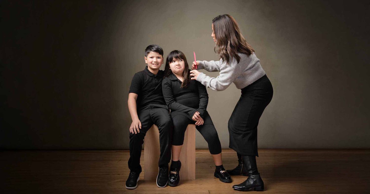 Two children in black outfits sit on wooden stools while a woman in a gray top styles one child's hair in a studio portrait.