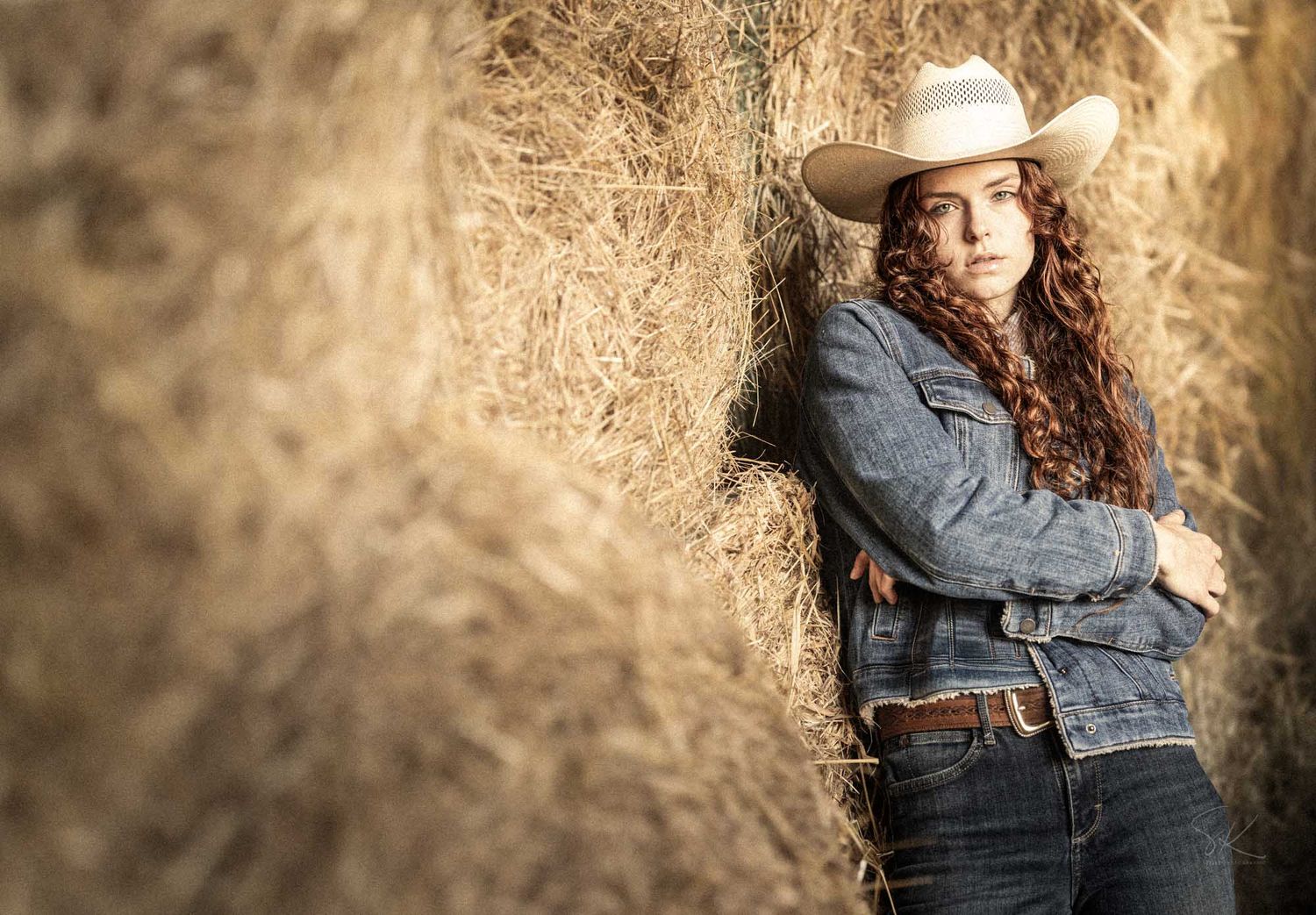 Woman in white cowboy hat and denim jacket leaning against hay bales with arms crossed.