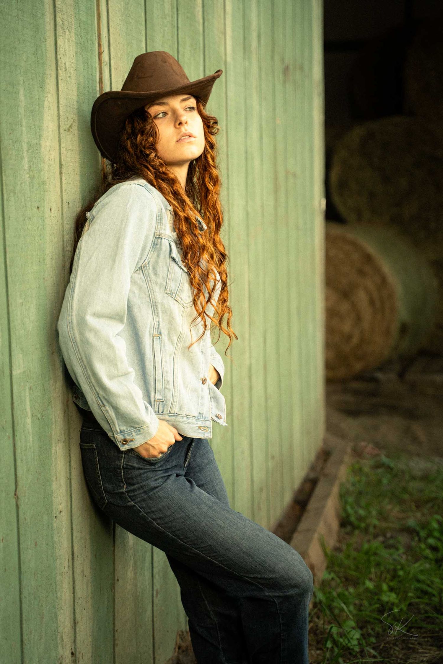 Young woman in cowboy hat and denim jacket leans against rustic barn wall with hay bales in background.