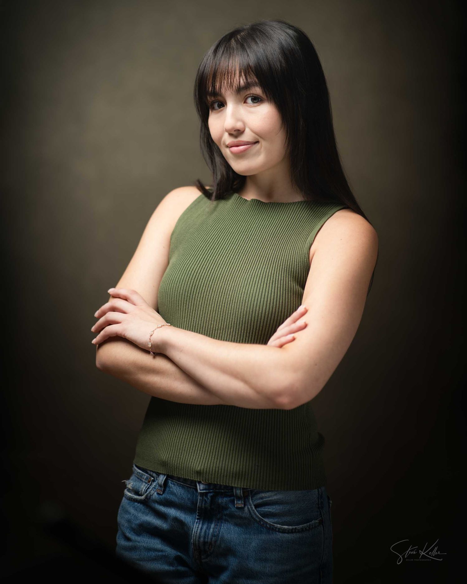 Young woman with dark hair in green sleeveless top and jeans, arms crossed, posing confidently against dark studio background.