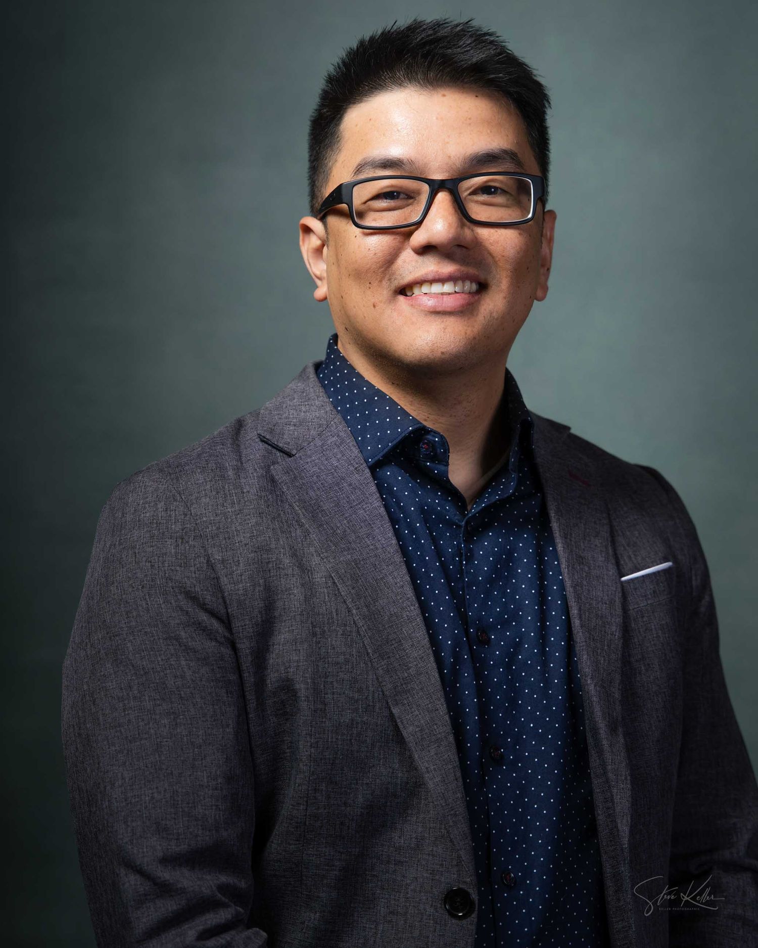 Smiling Asian man in gray blazer and navy dotted shirt wearing black glasses poses for professional headshot.