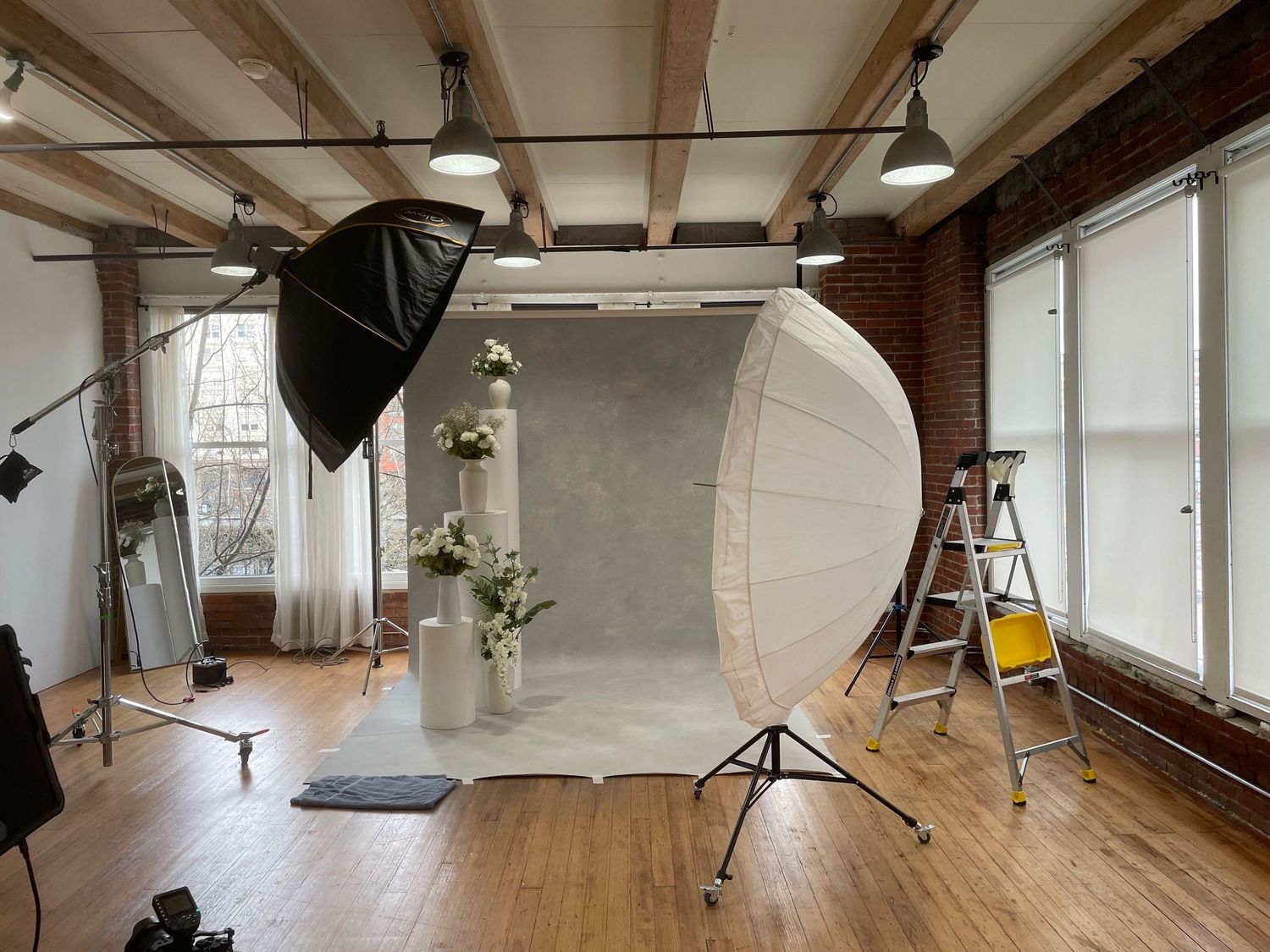 Professional photography studio with softbox lighting, large reflector, gray backdrop, floral arrangement, brick walls, and hardwood floors.