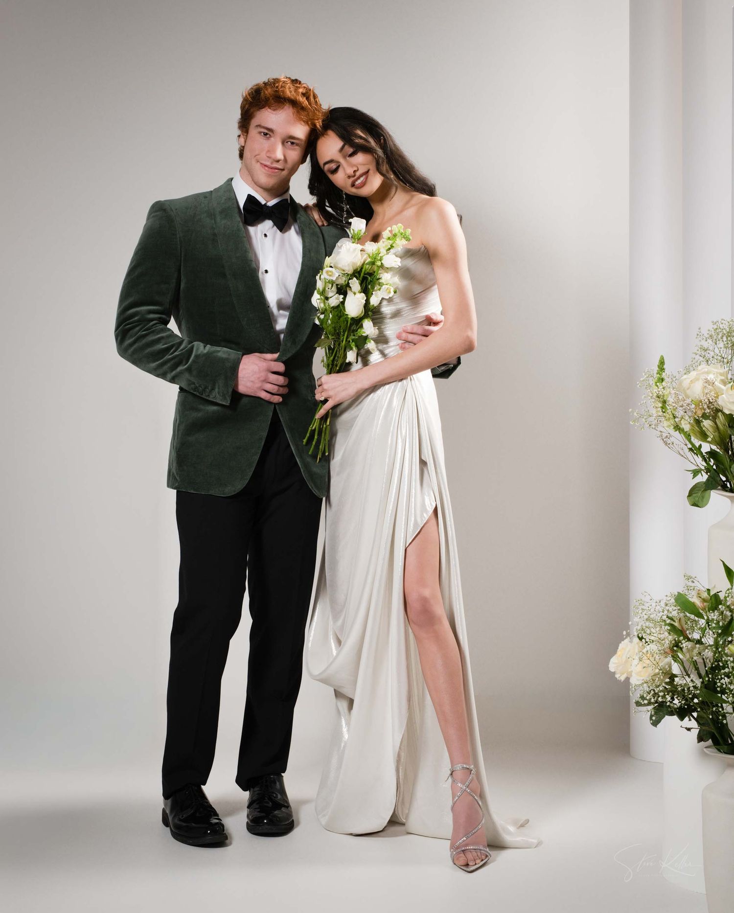 A couple in formal wedding attire pose together in a studio setting with white floral arrangements.