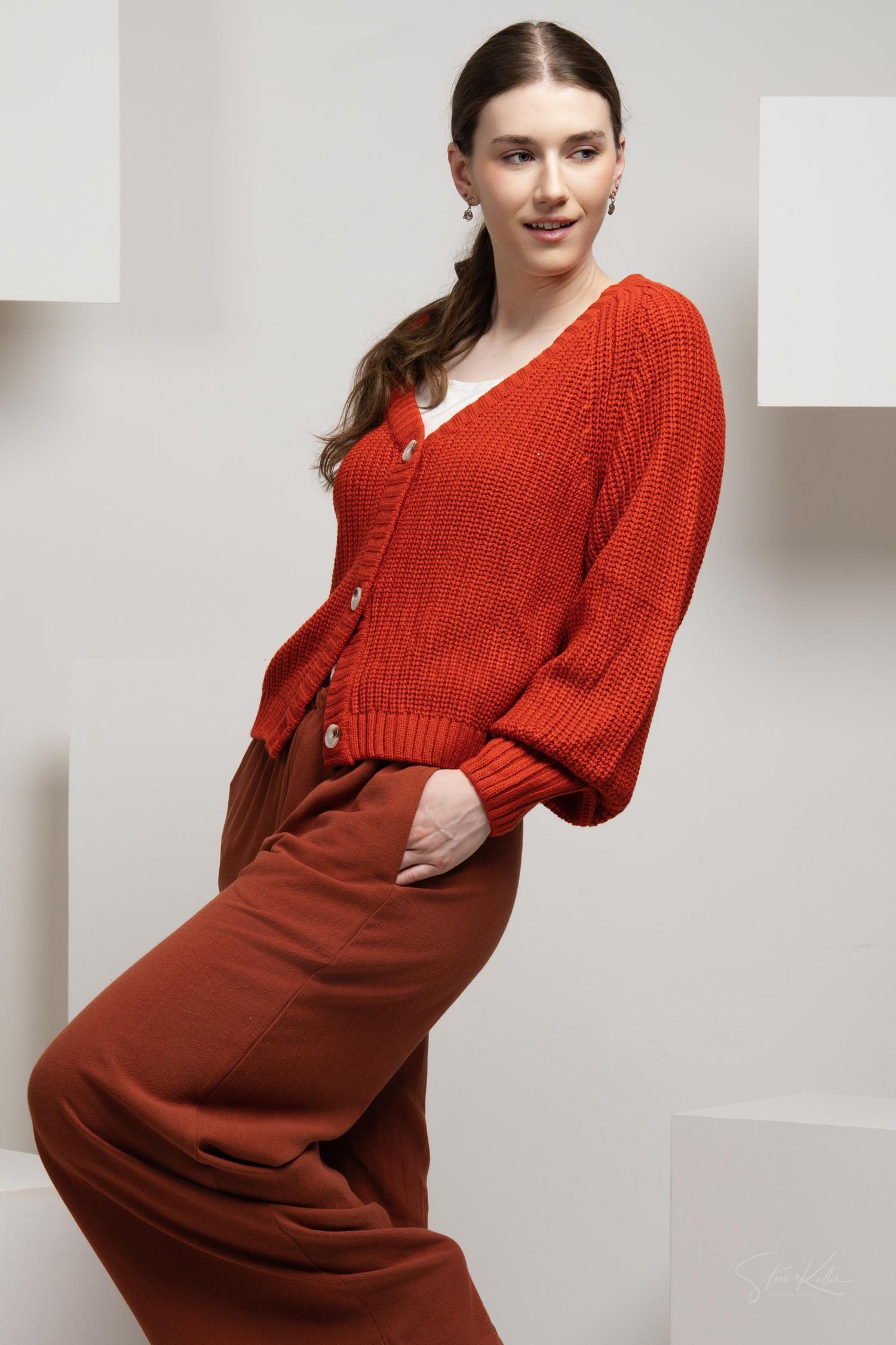 Woman in red knit cardigan and matching pants poses elegantly against white geometric backdrop.