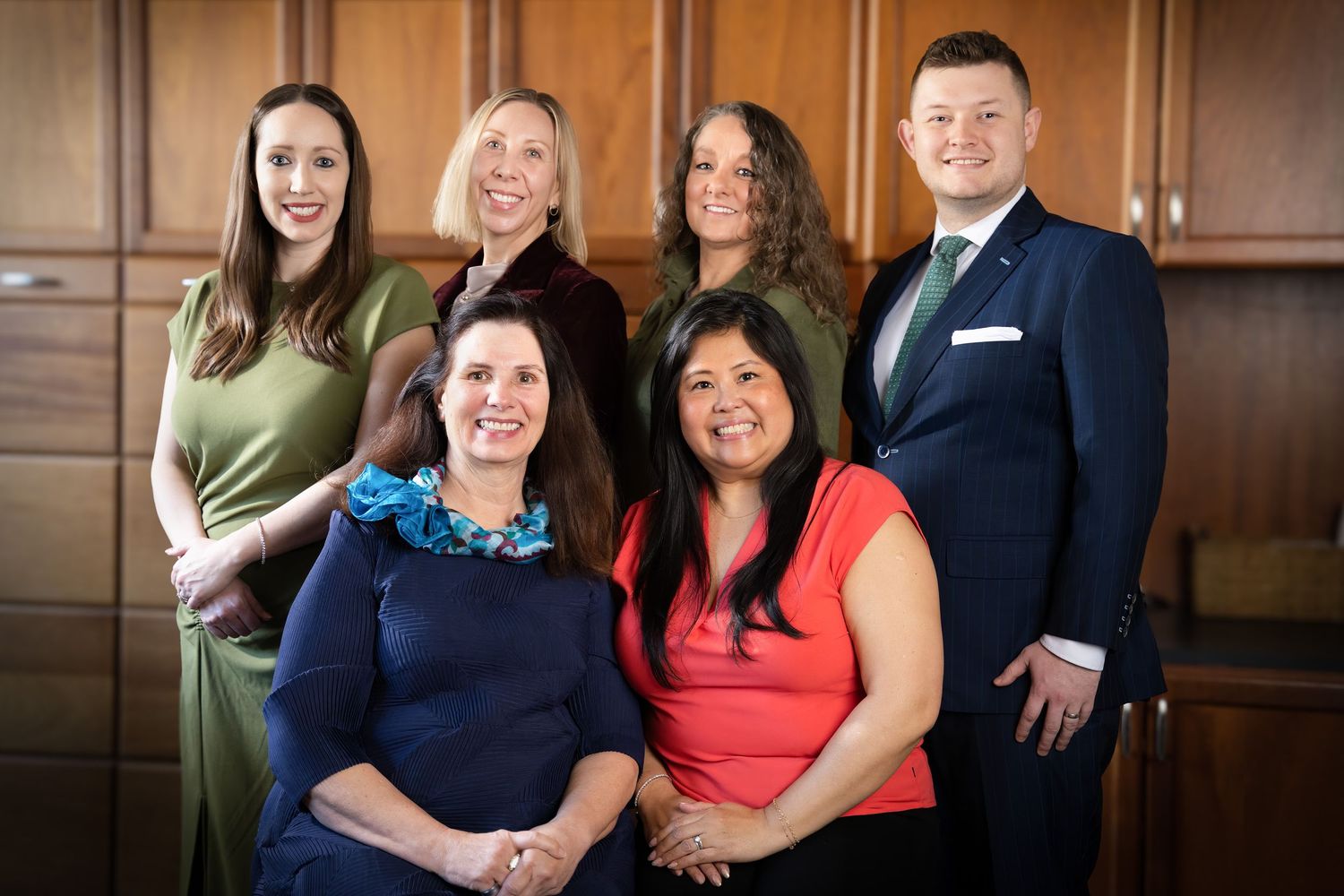 Six professional colleagues pose together smiling in a formal office setting, wearing business attire.