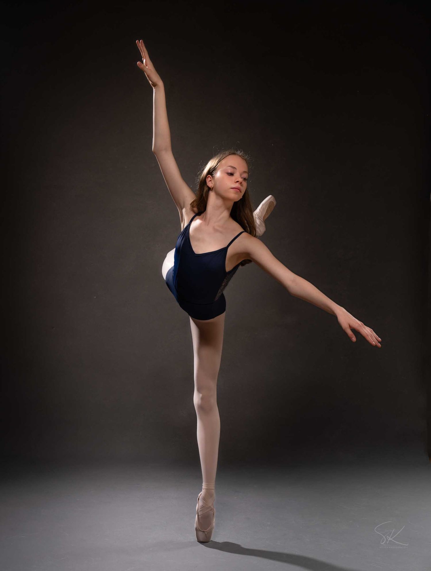 Young ballet dancer in navy leotard and pointe shoes performs an arabesque against a dark studio background.