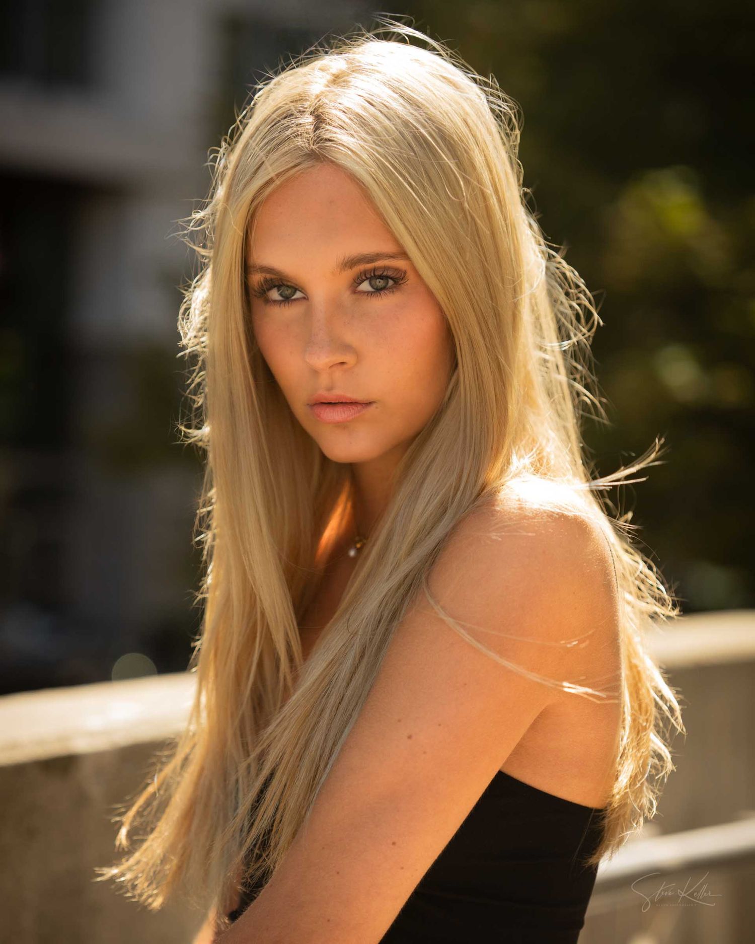 Young blonde woman with long straight hair poses outdoors in sunlight wearing a black top.