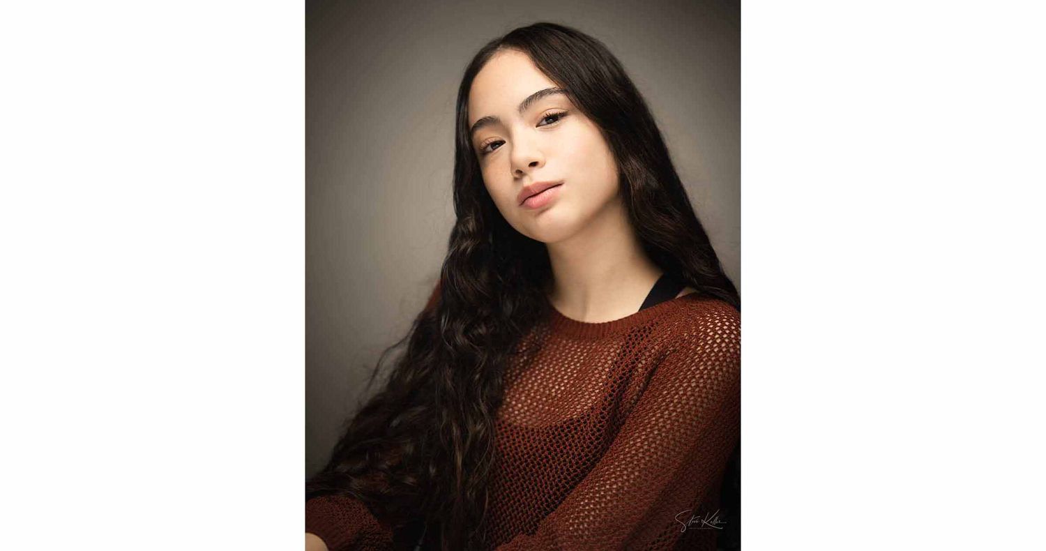 Young woman with long dark wavy hair wearing a rust-brown knit top poses for a studio portrait against a neutral gray backdrop.