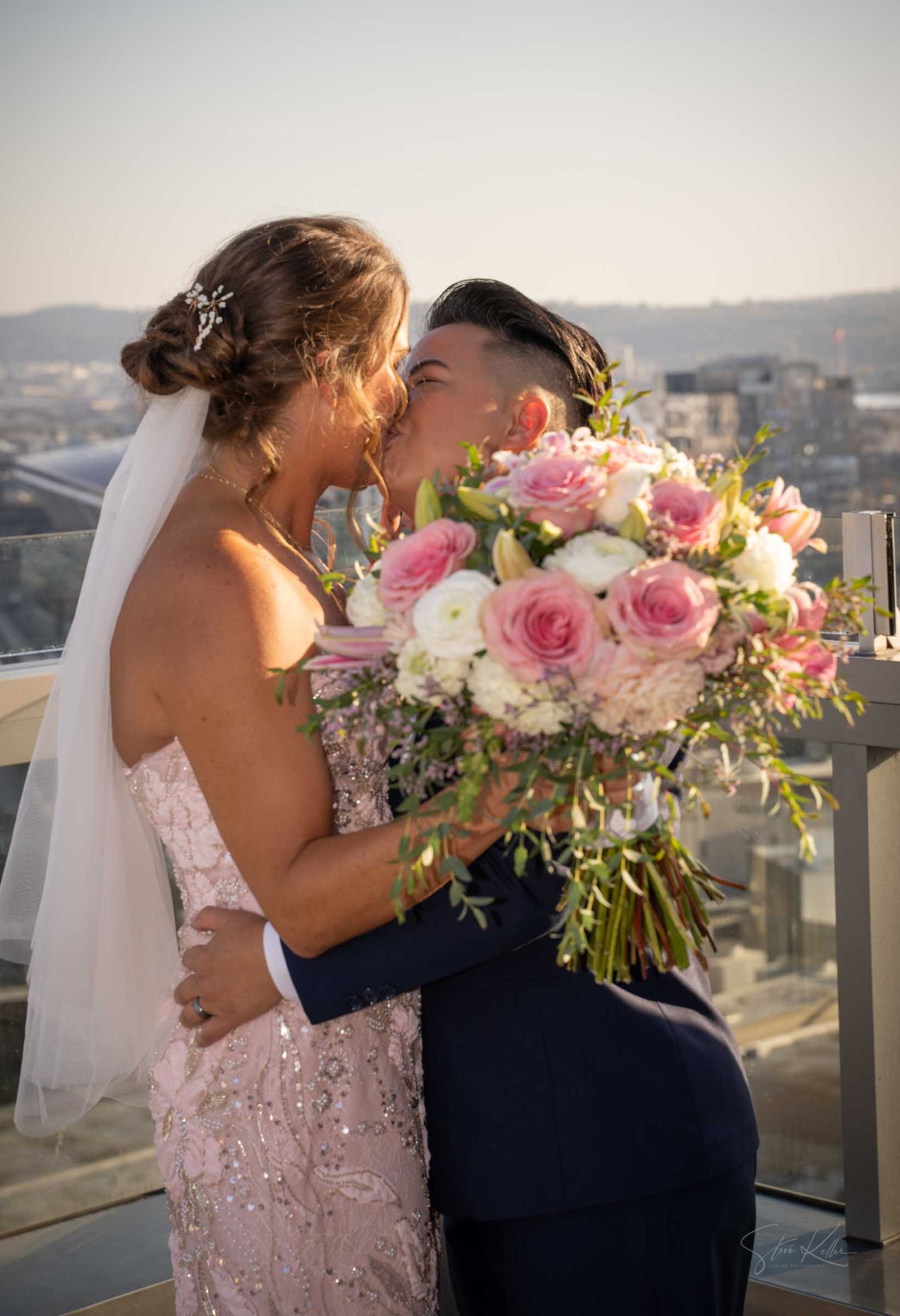 Two brides kissing on a rooftop venue holding a pink and white rose bouquet with city skyline views.