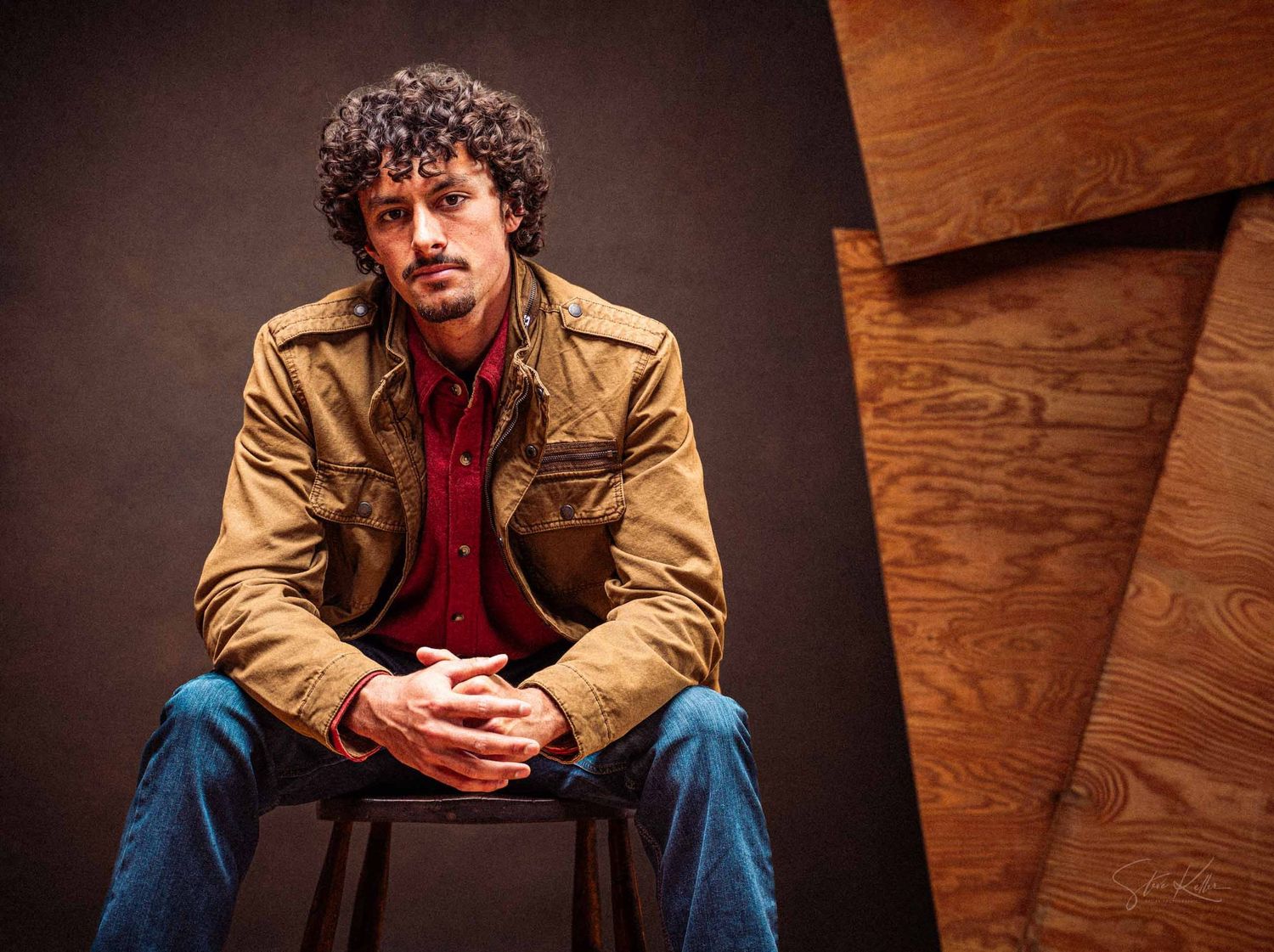 Person in tan jacket and red shirt sits on wooden floor in multiple candid poses.