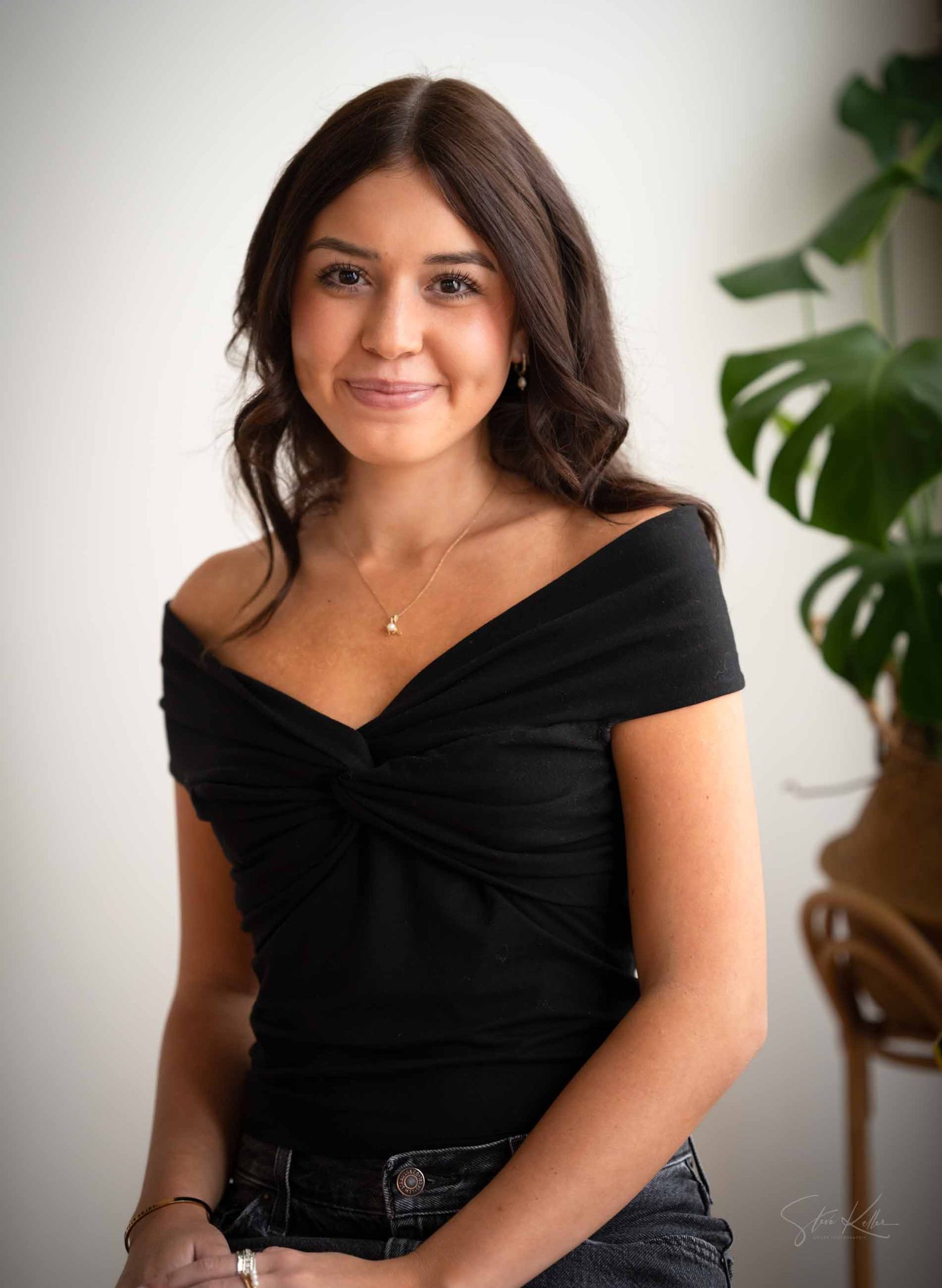 Woman in black off-shoulder top poses against white wall with indoor plant in background.