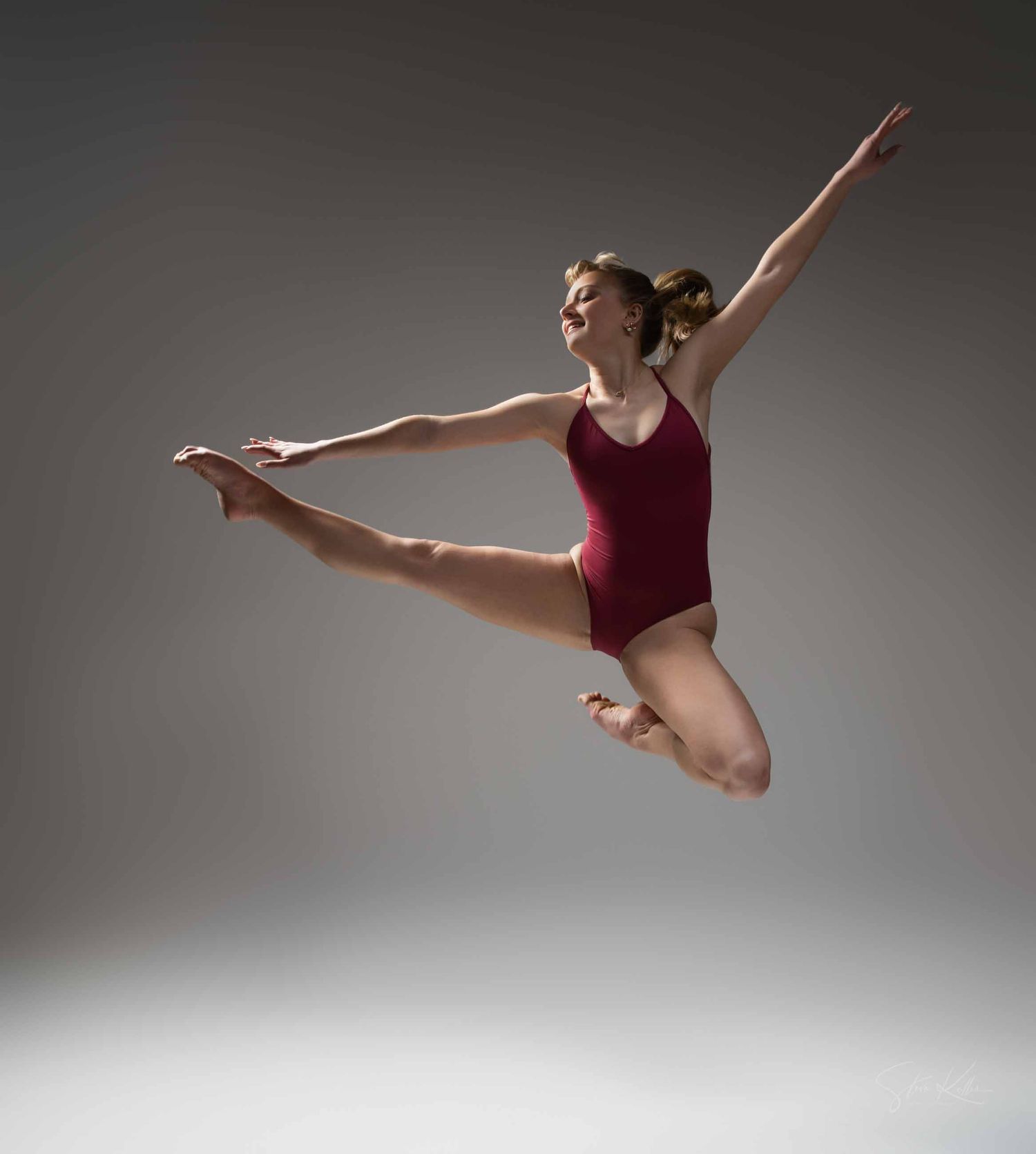 Dancer in burgundy leotard performs dramatic leap against neutral studio background.