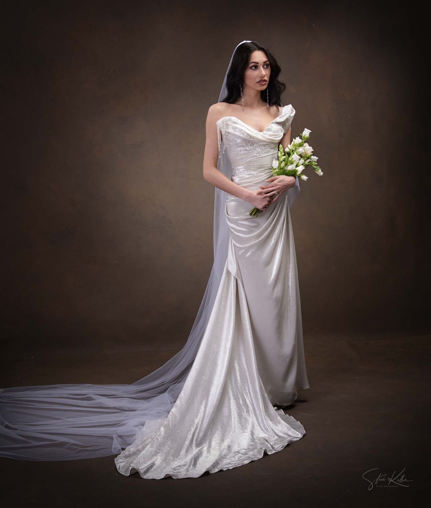 A bride in a flowing white satin wedding gown with a long train poses elegantly against a dark studio backdrop.